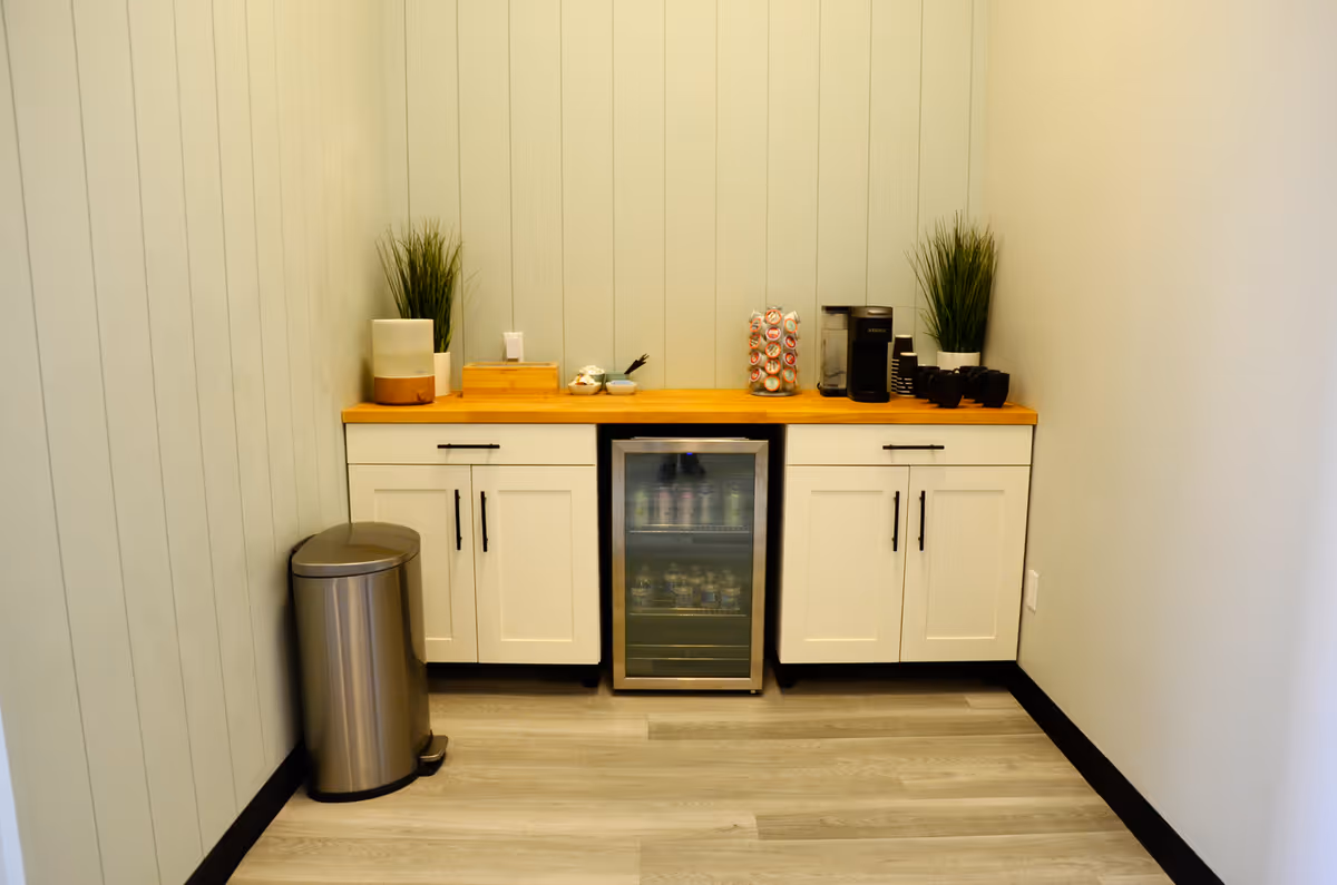 Small coffee station with white cabinets, a mini-fridge, and plants on a wooden counter.