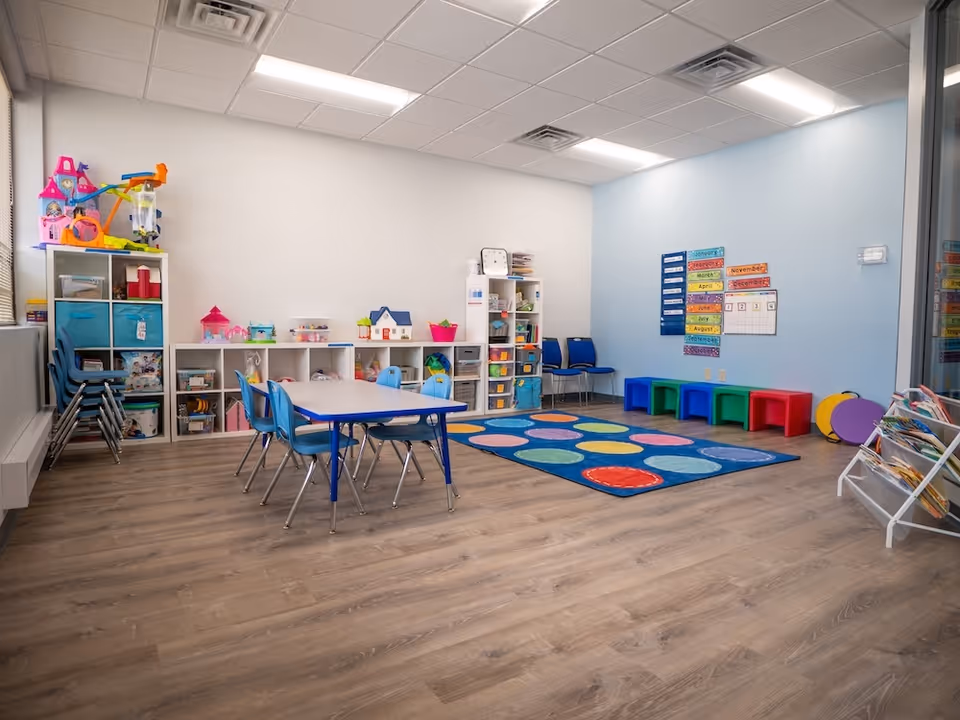 Bright children's playroom with tables, chairs, shelves, and a colorful circular rug.