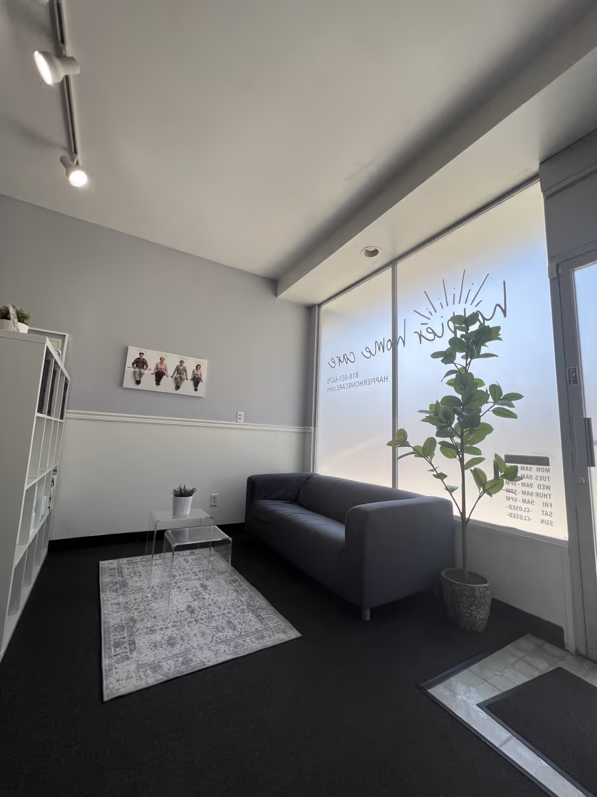 Facility lobby lounge with gray sofa, glass coffee tables, rug, plant, and frosted windows.