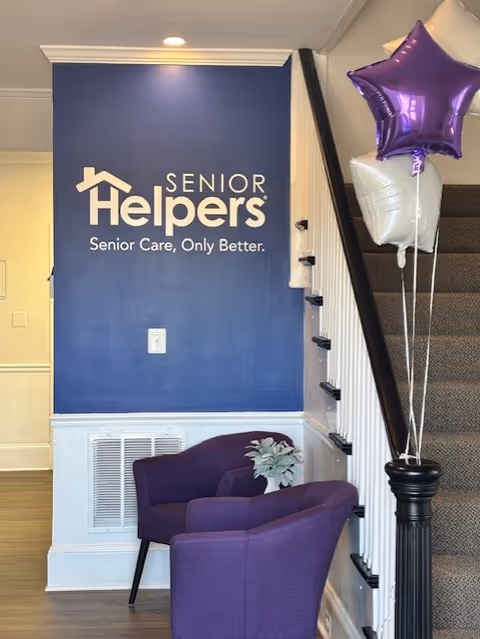 Blue wall with Senior Helpers logo, purple chairs, and balloons near a stairway in a lobby.