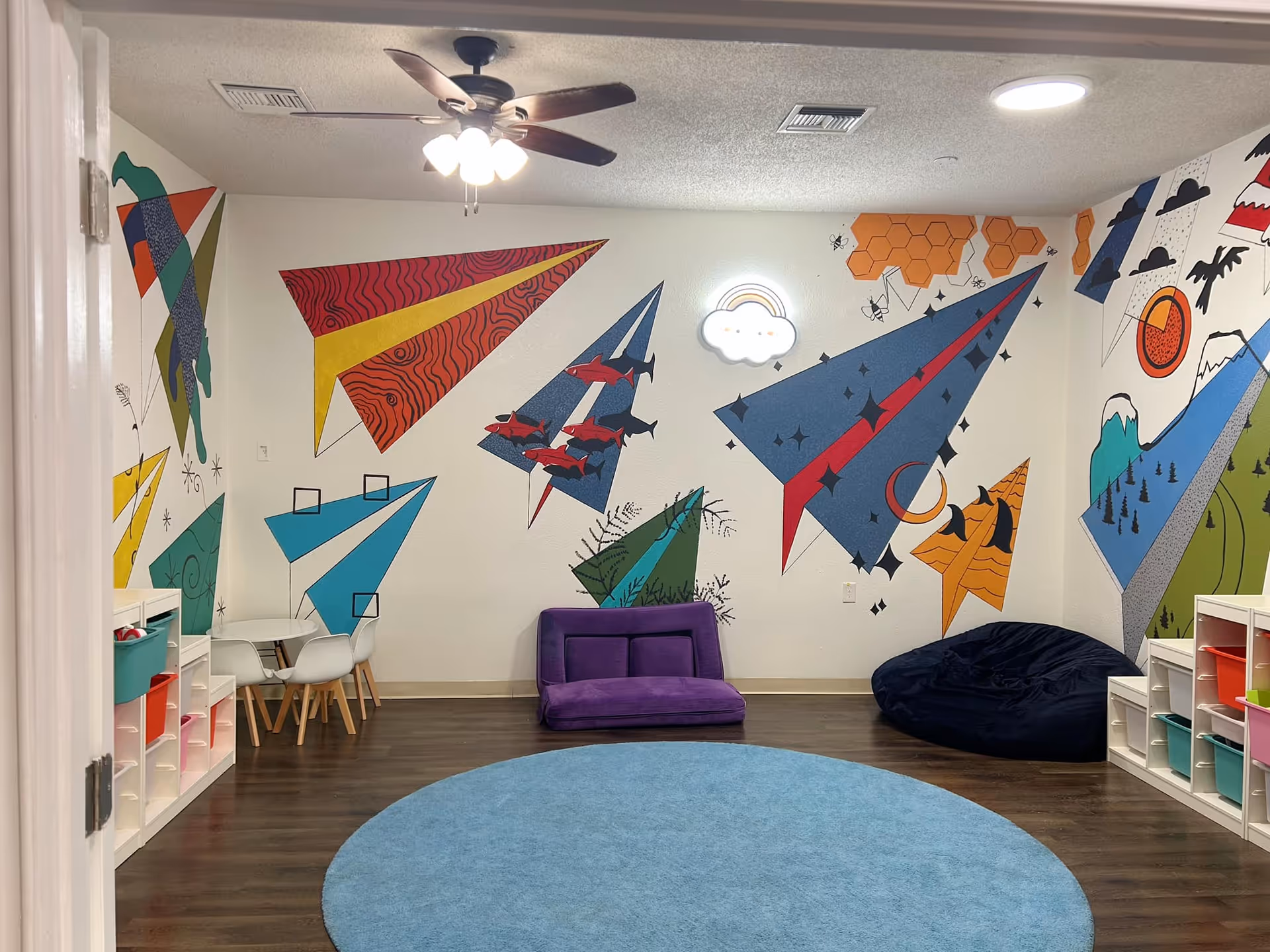 Colorful geometric murals brighten a children's play area with seating, shelves and a blue circular rug.
