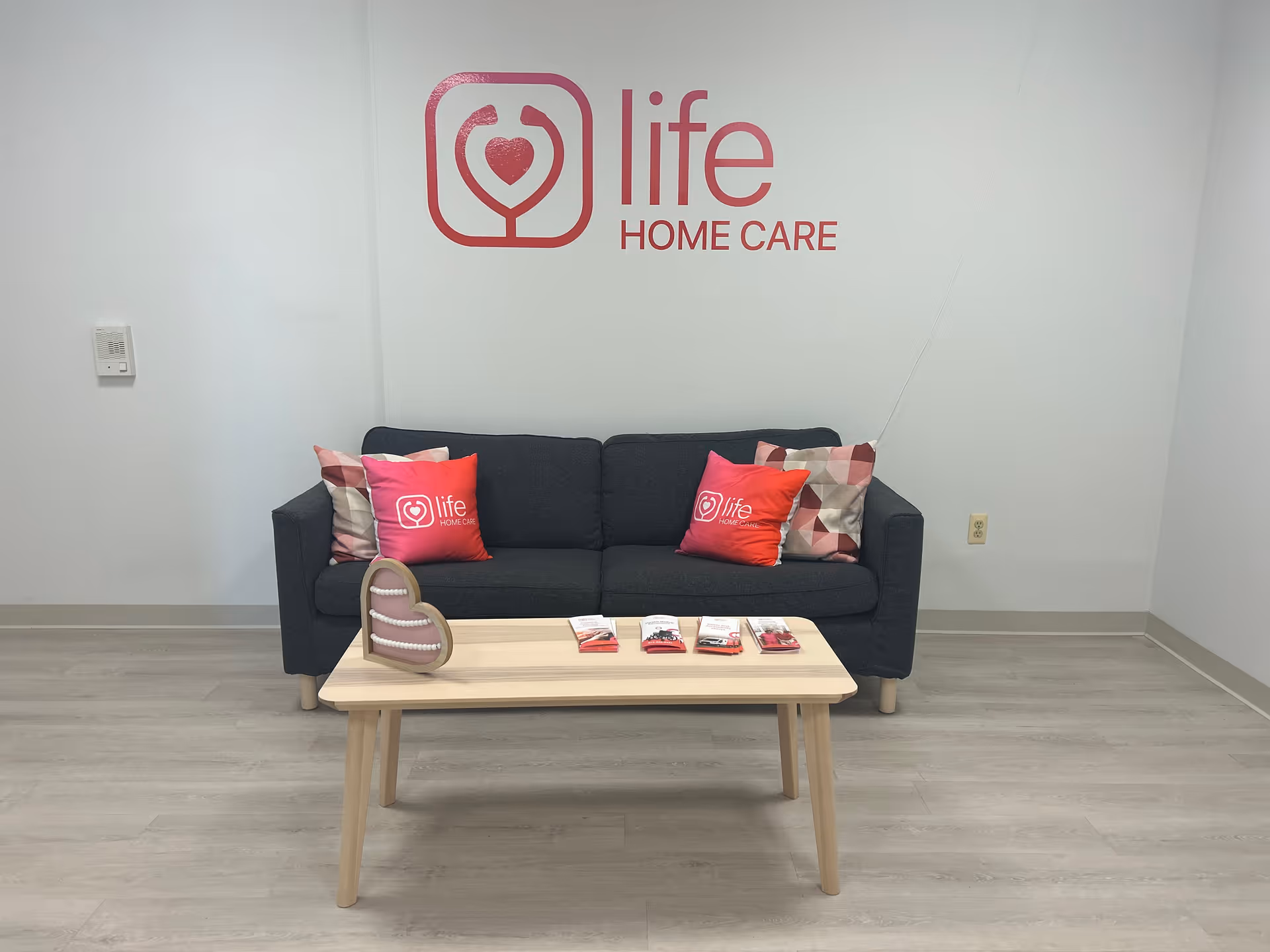 A waiting area with a dark sofa, branded red pillows, and a Life Home Care logo.