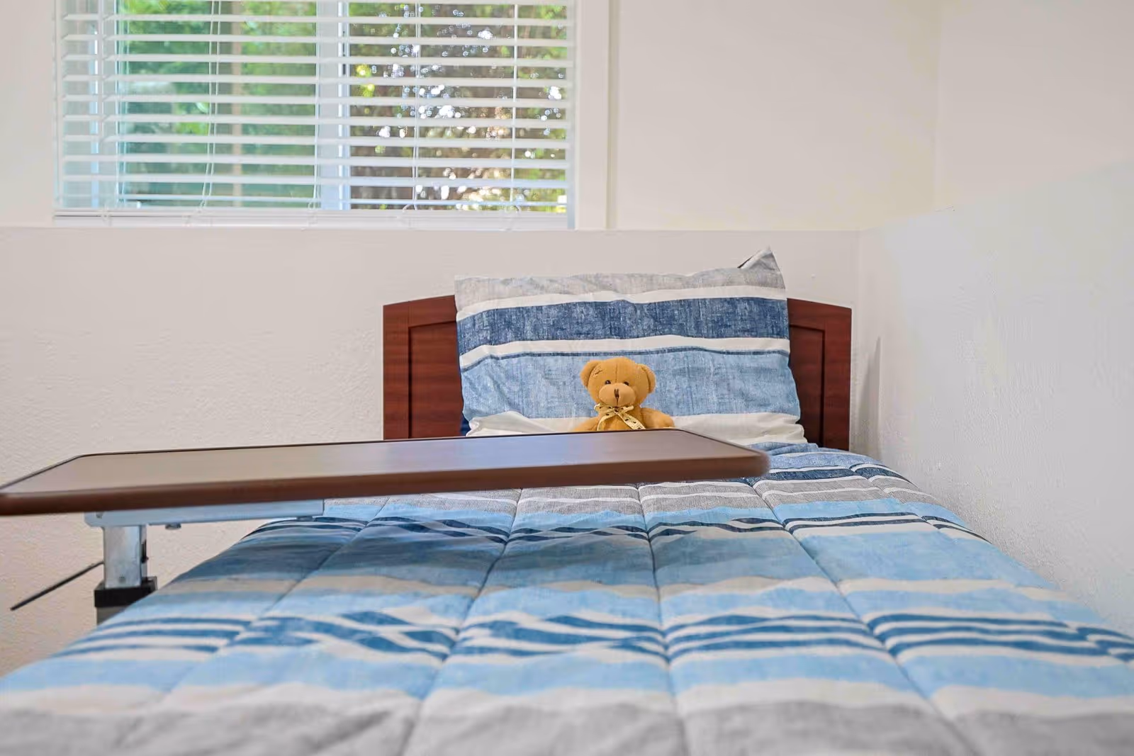 Bedroom with blue striped bedding, overbed table, a teddy bear, and window blinds.