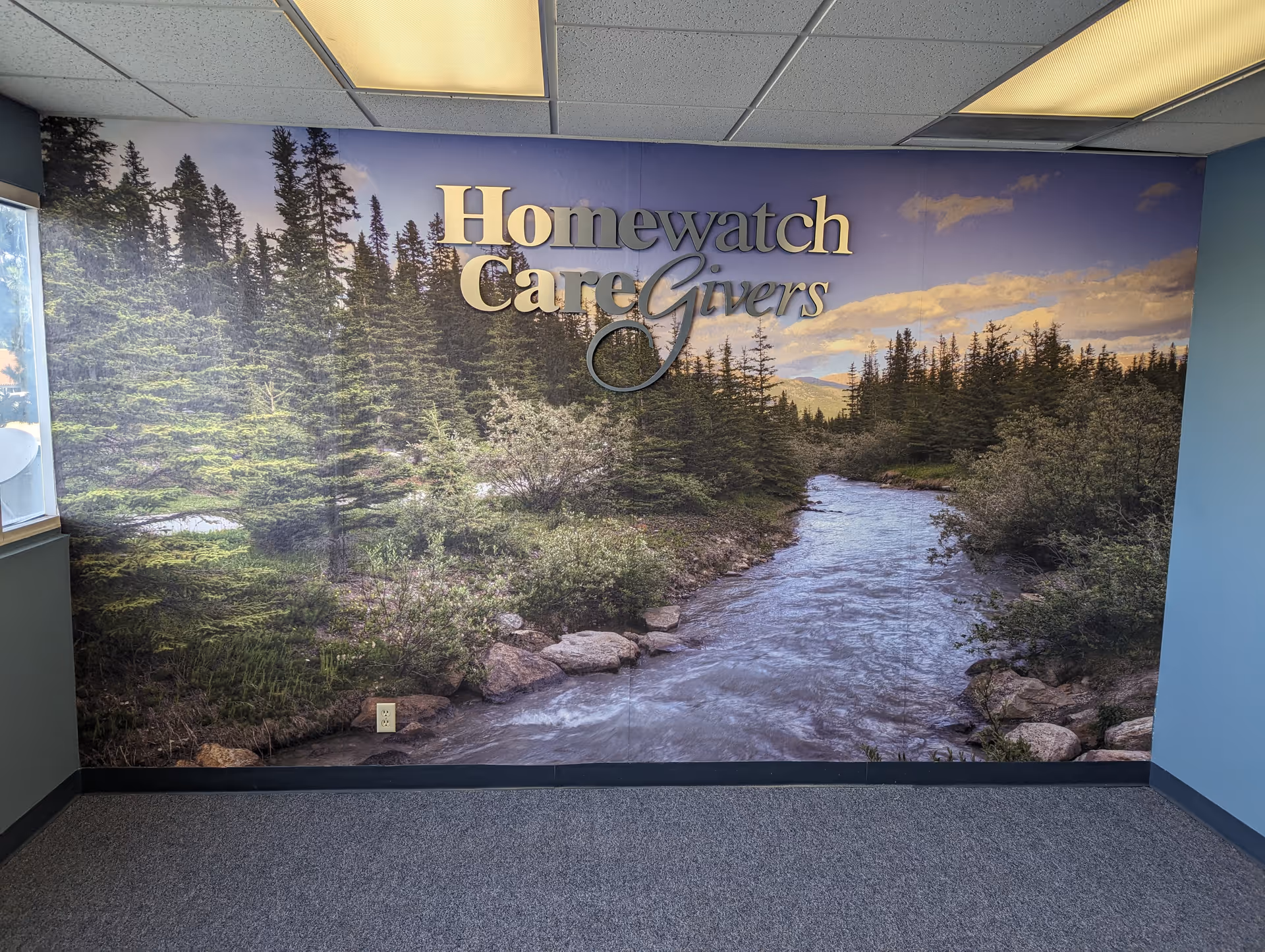 Interior hallway wall features a large mural of a river and trees with Homewatch CareGivers logo.