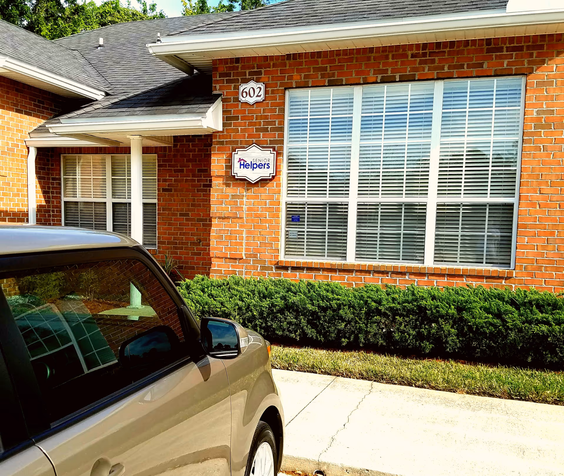 Brick senior living building front with large windows, a car, and Senior Helpers sign.