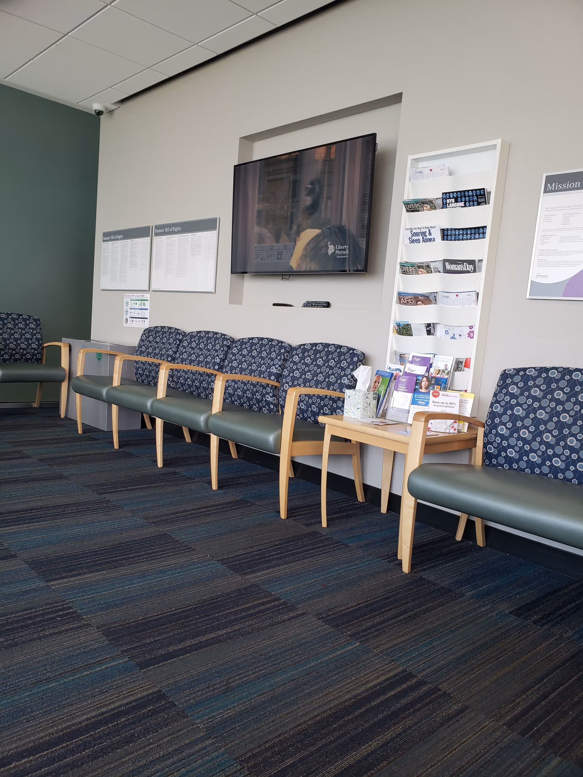 A row of blue upholstered chairs lines the wall with a mounted TV and pamphlet rack.