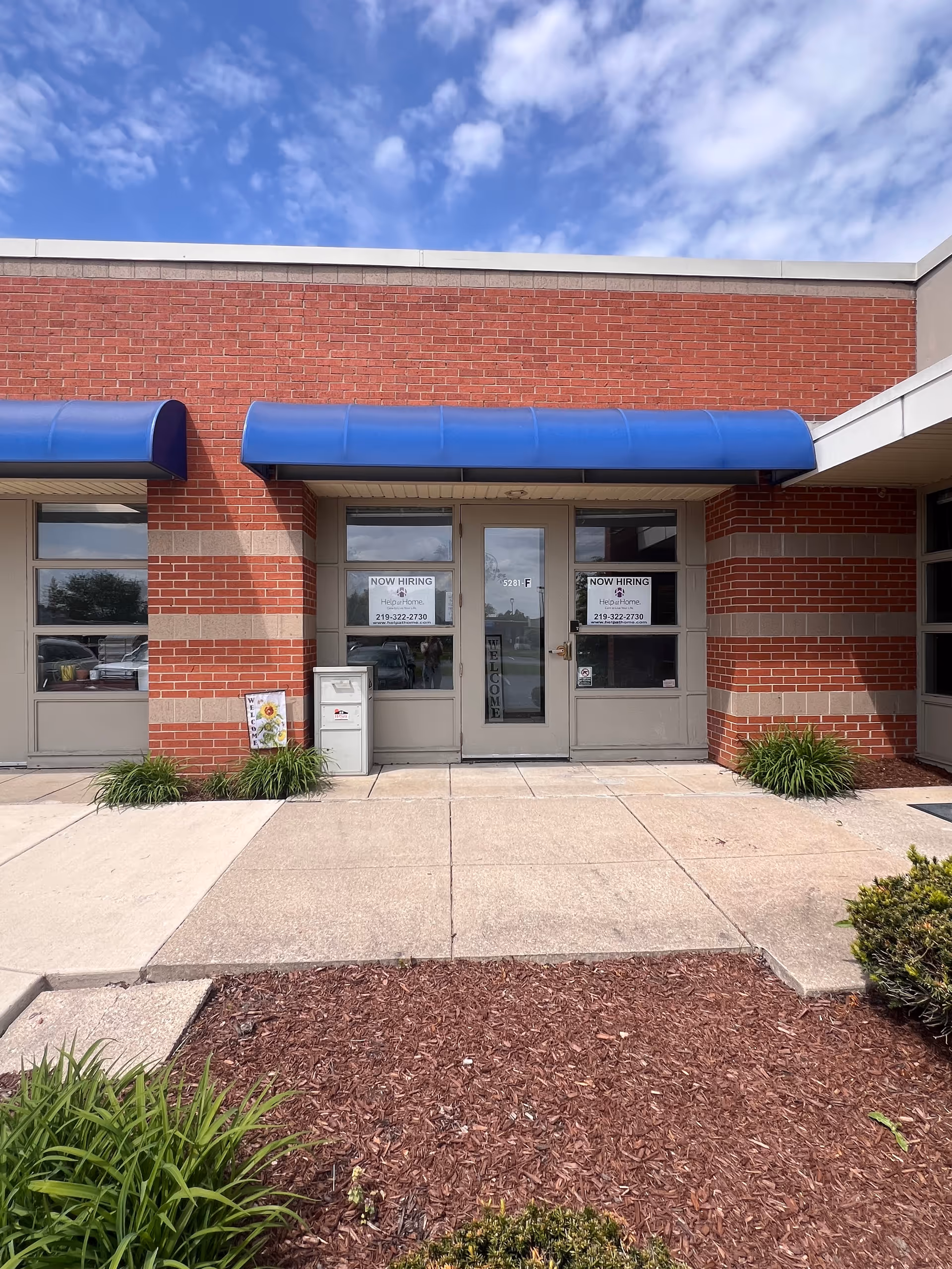 Brick facility entrance with blue awnings, glass doors, and Now Hiring signs.