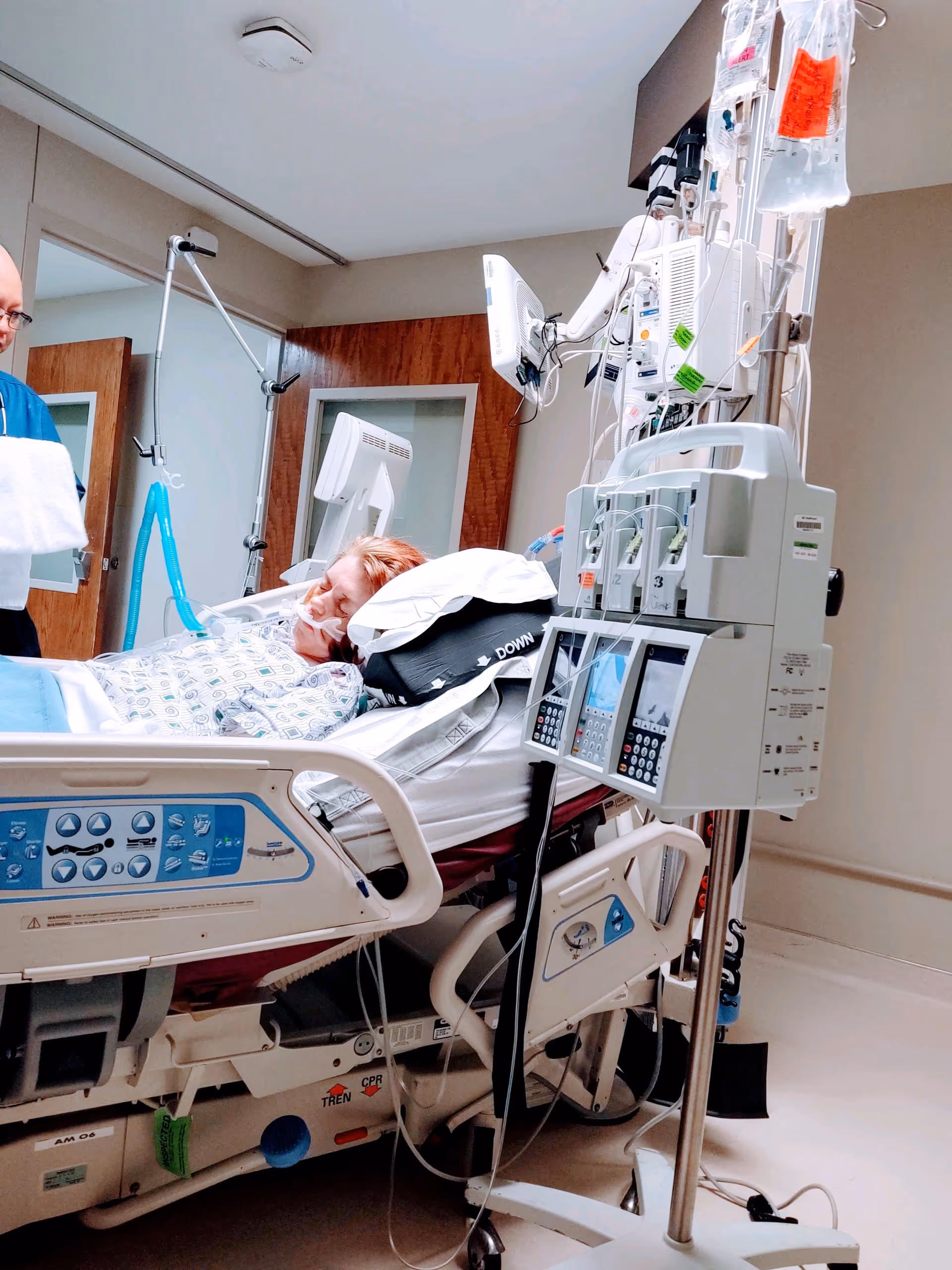 Patient in hospital bed connected to monitors and IVs with a caregiver nearby.