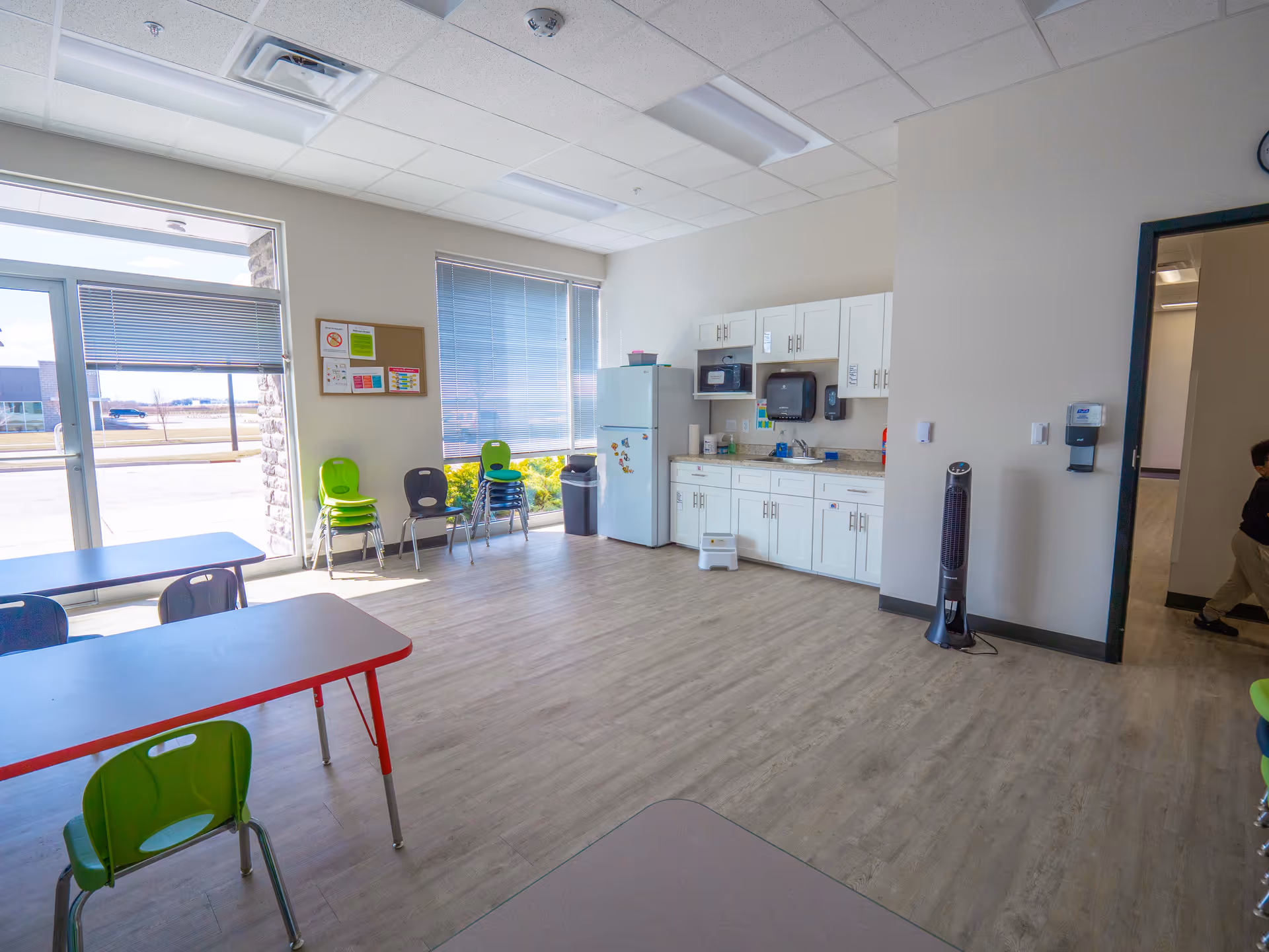 Bright communal kitchen and dining area with tables, chairs, and appliances inside a facility.