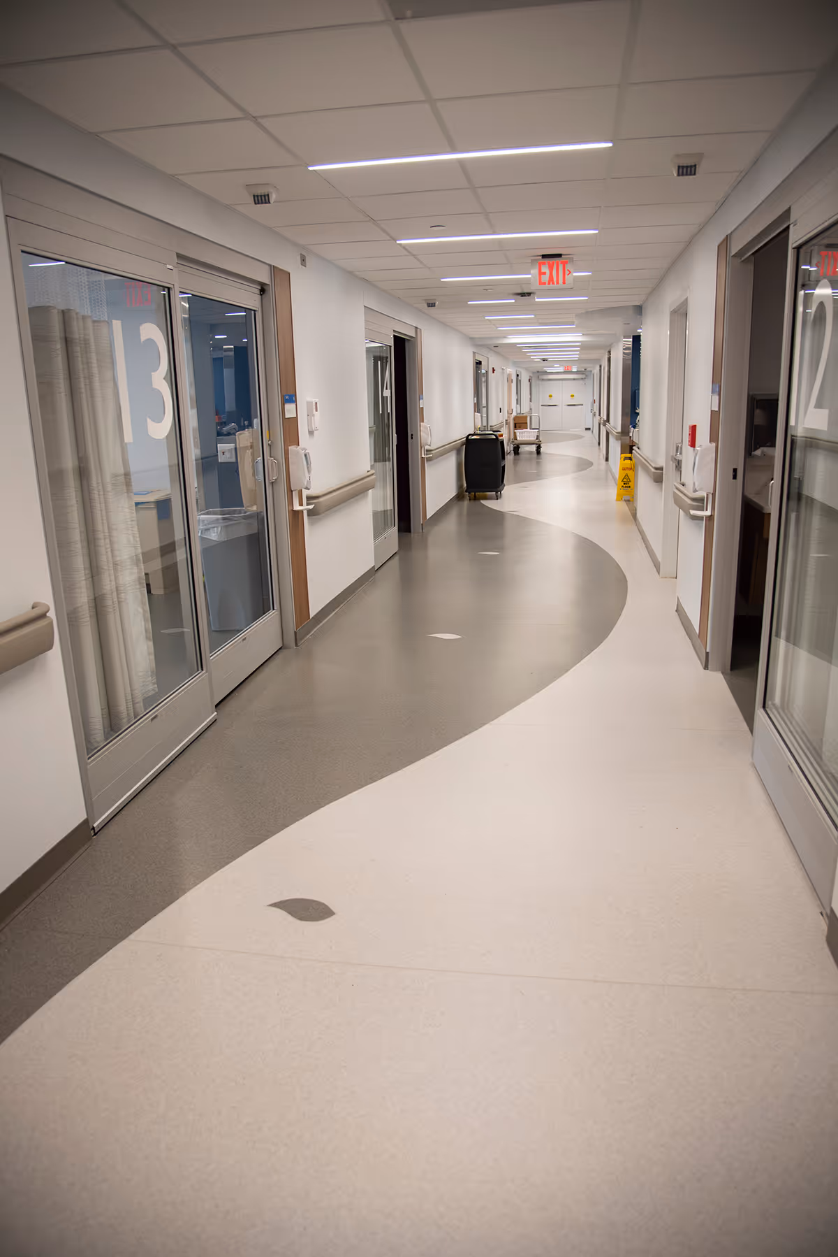 Bright hospital corridor with numbered rooms, glass doors, and an exit sign along the hall.