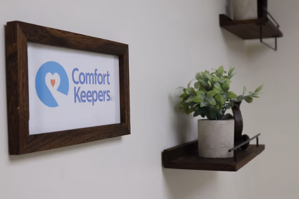 Interior wall features a Comfort Keepers sign framed in wood beside a plant.