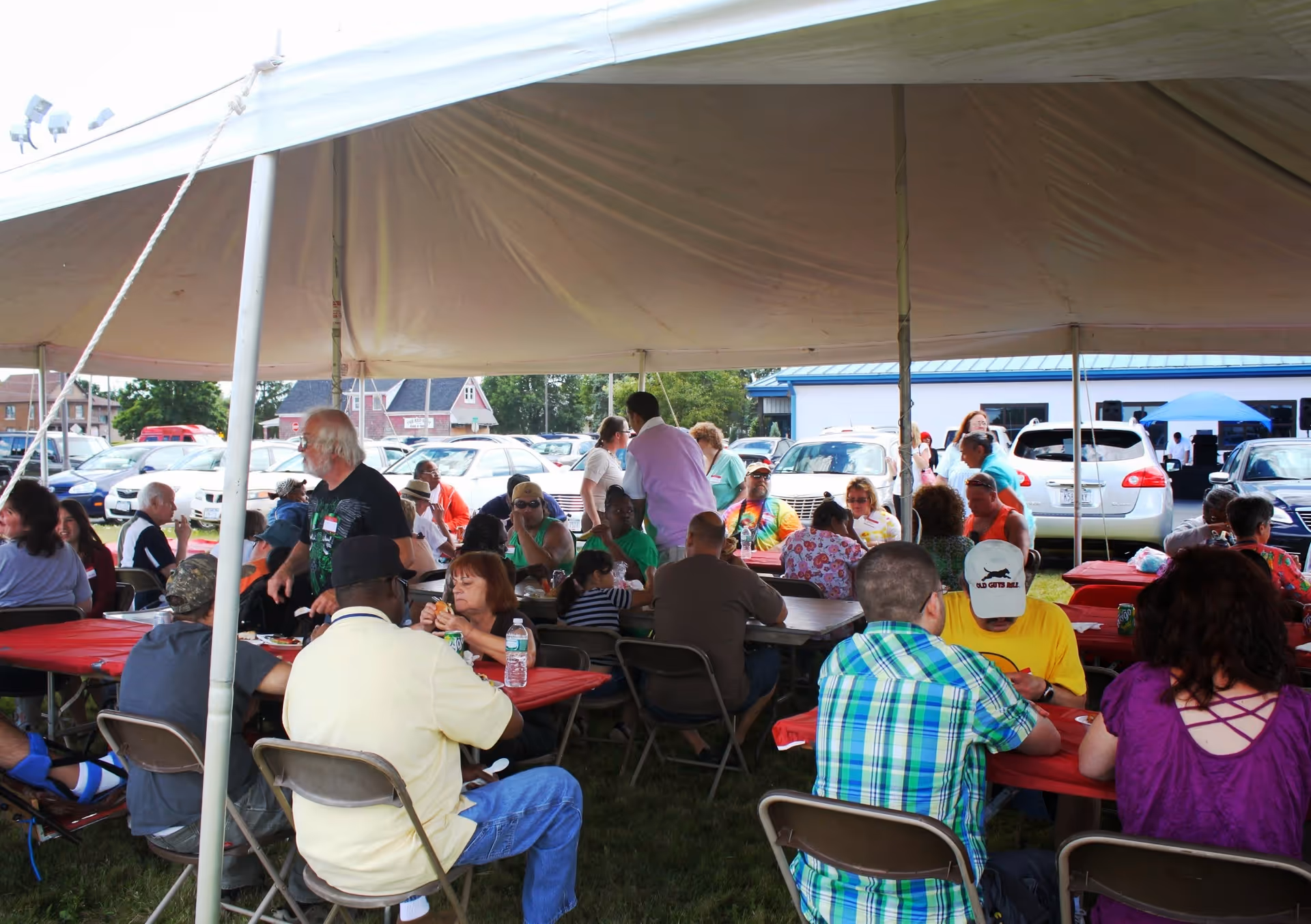 Outdoor tented gathering with numerous people seated at red-tablecloth tables.