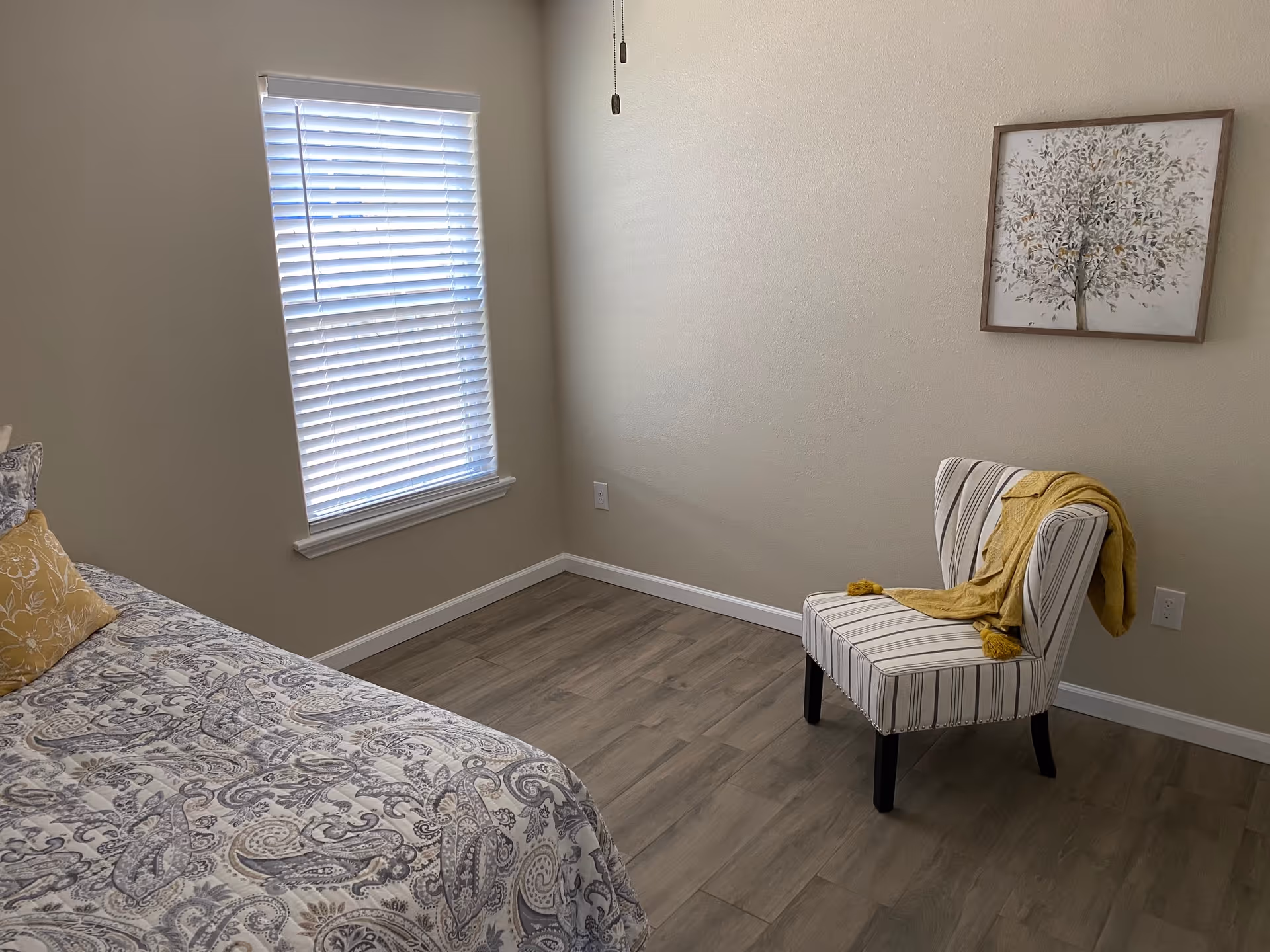 A cozy bedroom with a bed, striped chair, wall art, and window blinds.