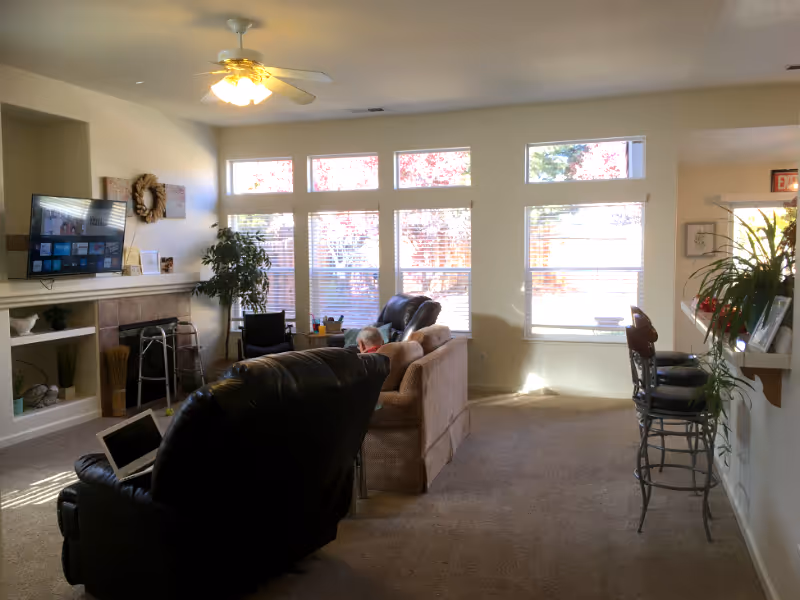 Sunlit senior living room with comfortable seating, a fireplace, and large windows.