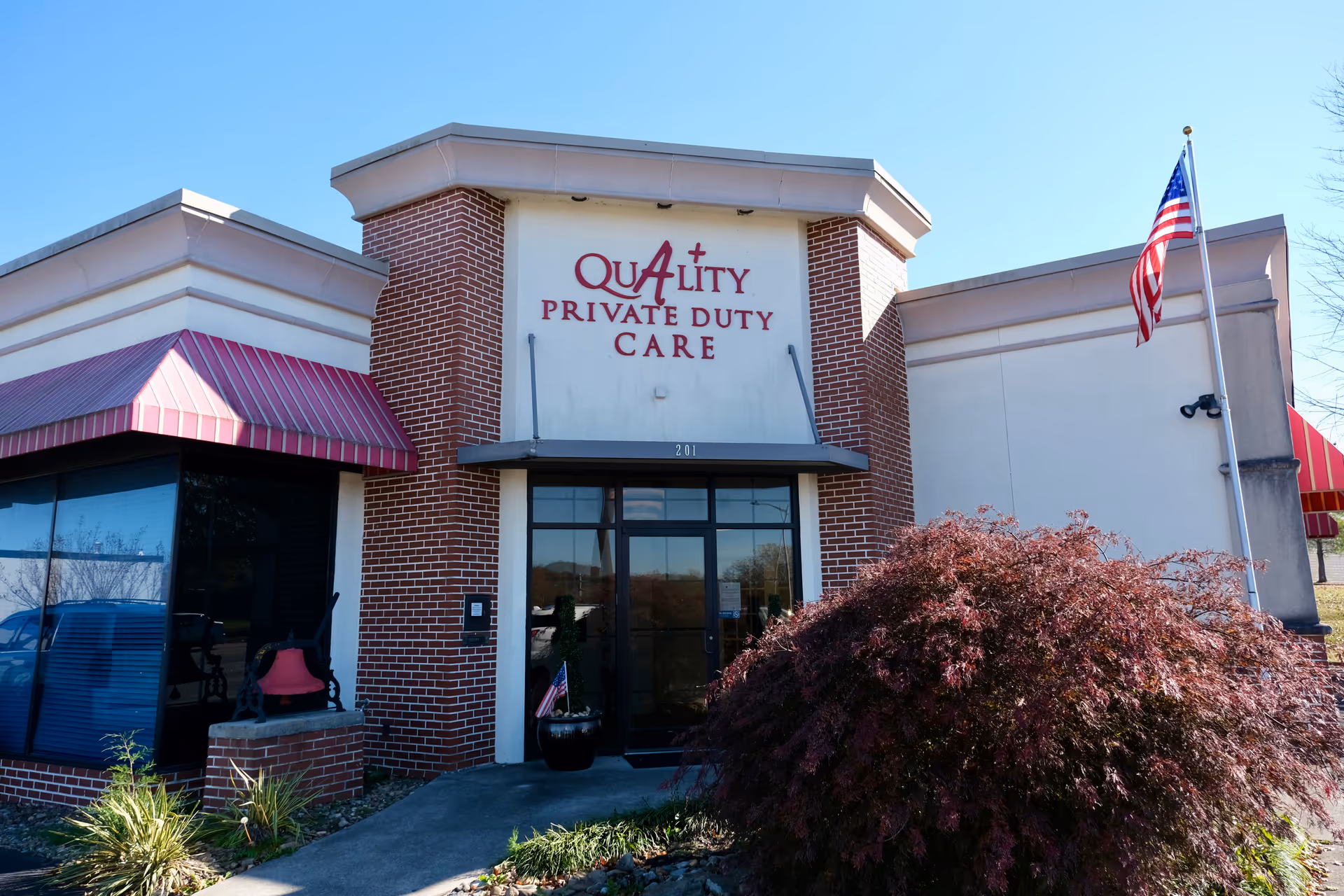 Front view of a brick facility entrance with Quality Private Duty Care sign and American flag.
