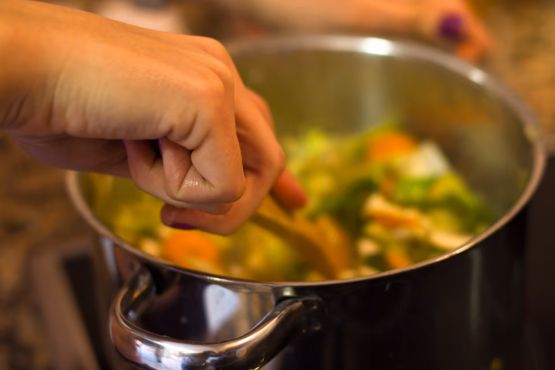 A hand stirs a pot of vegetables on a stovetop.