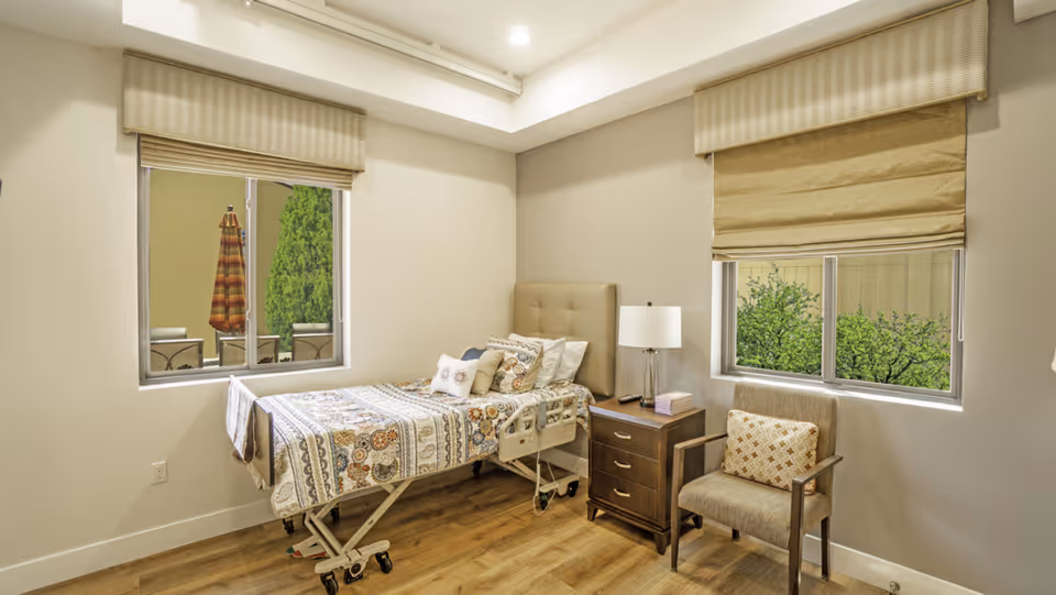 A neutral bedroom with a hospital-style bed, nightstand, chair, and two windows with blinds.