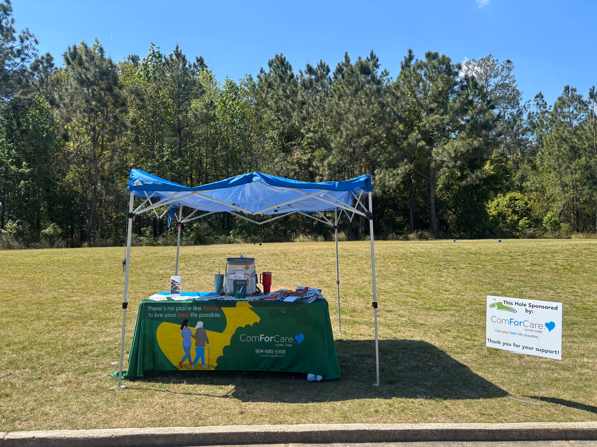 Outdoor event booth with a ComForCare table under a blue canopy in a grassy field