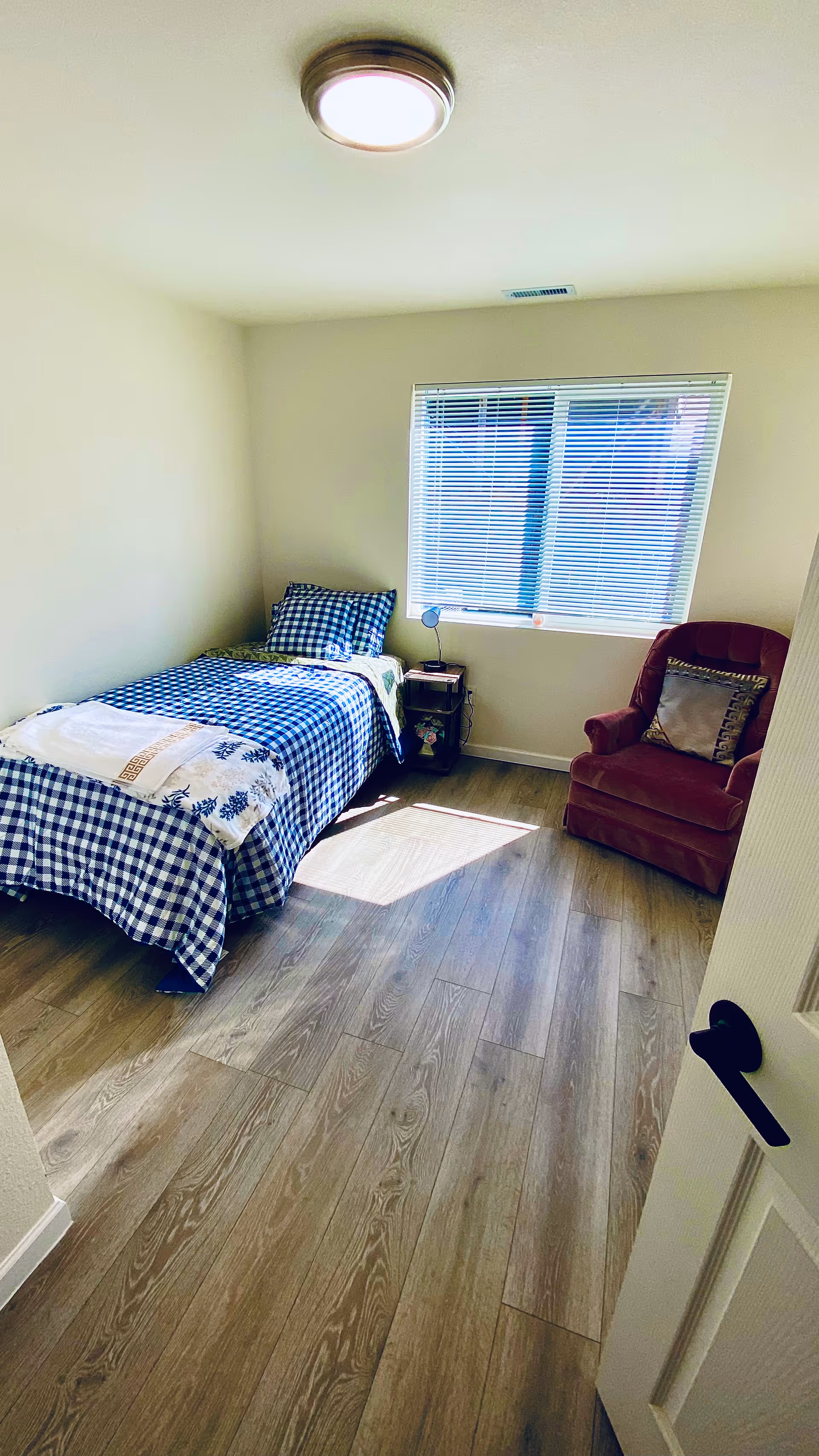 Small bedroom with twin bed, blue checkered bedding, window, and a red armchair.