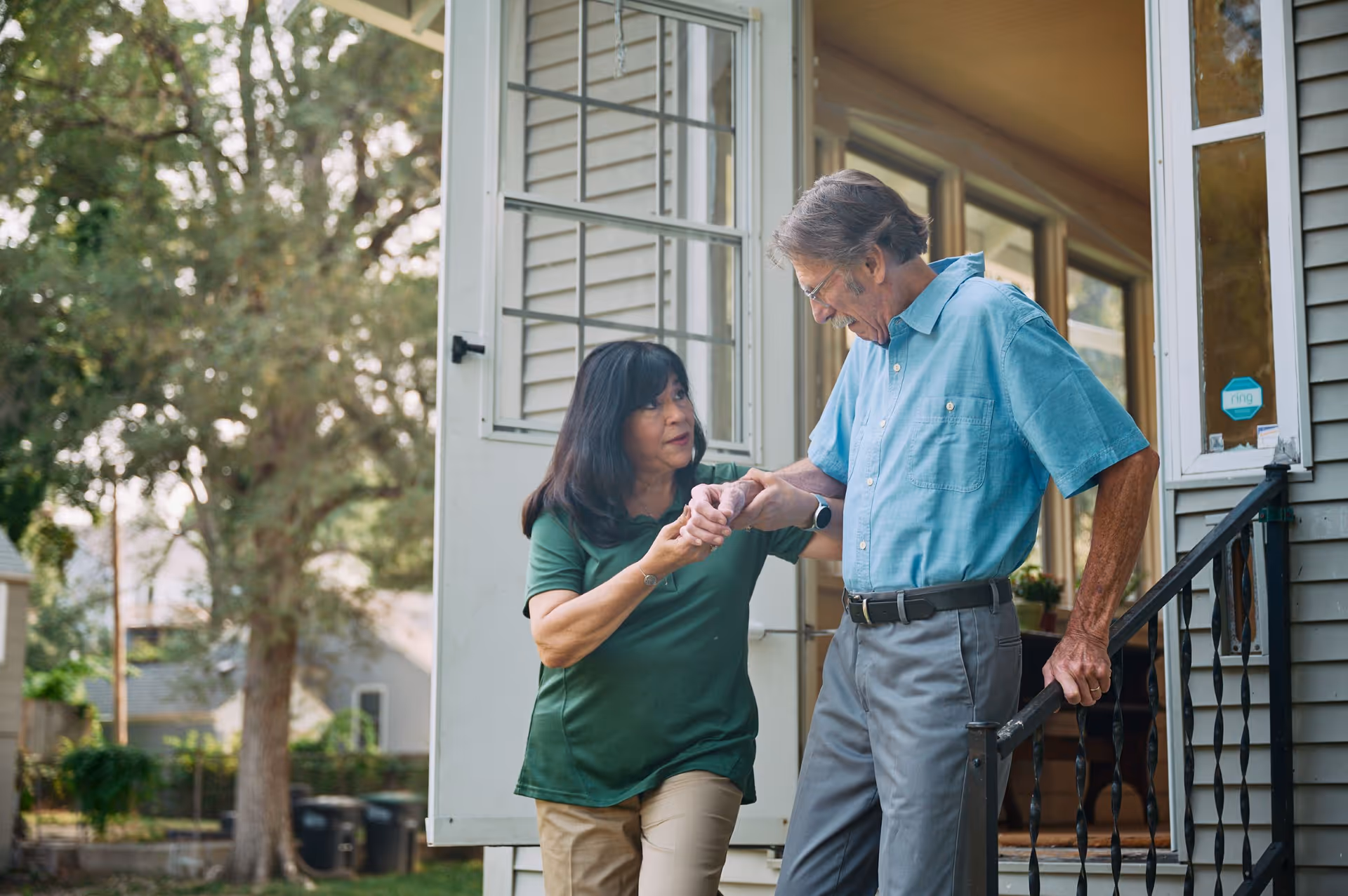 A caregiver assists an elderly man on outdoor steps at a home entrance.