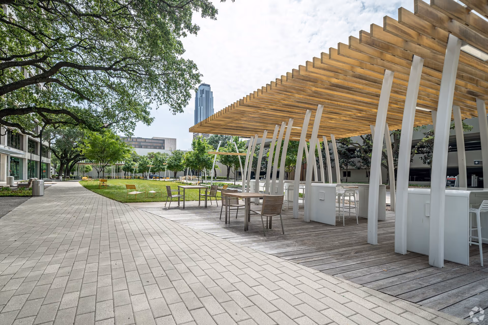 Outdoor courtyard with wooden pergola, seating, and trees beside a modern building.