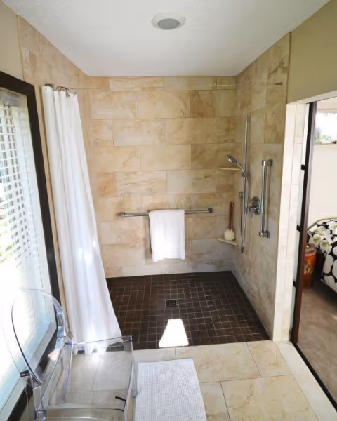 A walk-in tiled shower in a beige bathroom with a towel bar and shower chair.