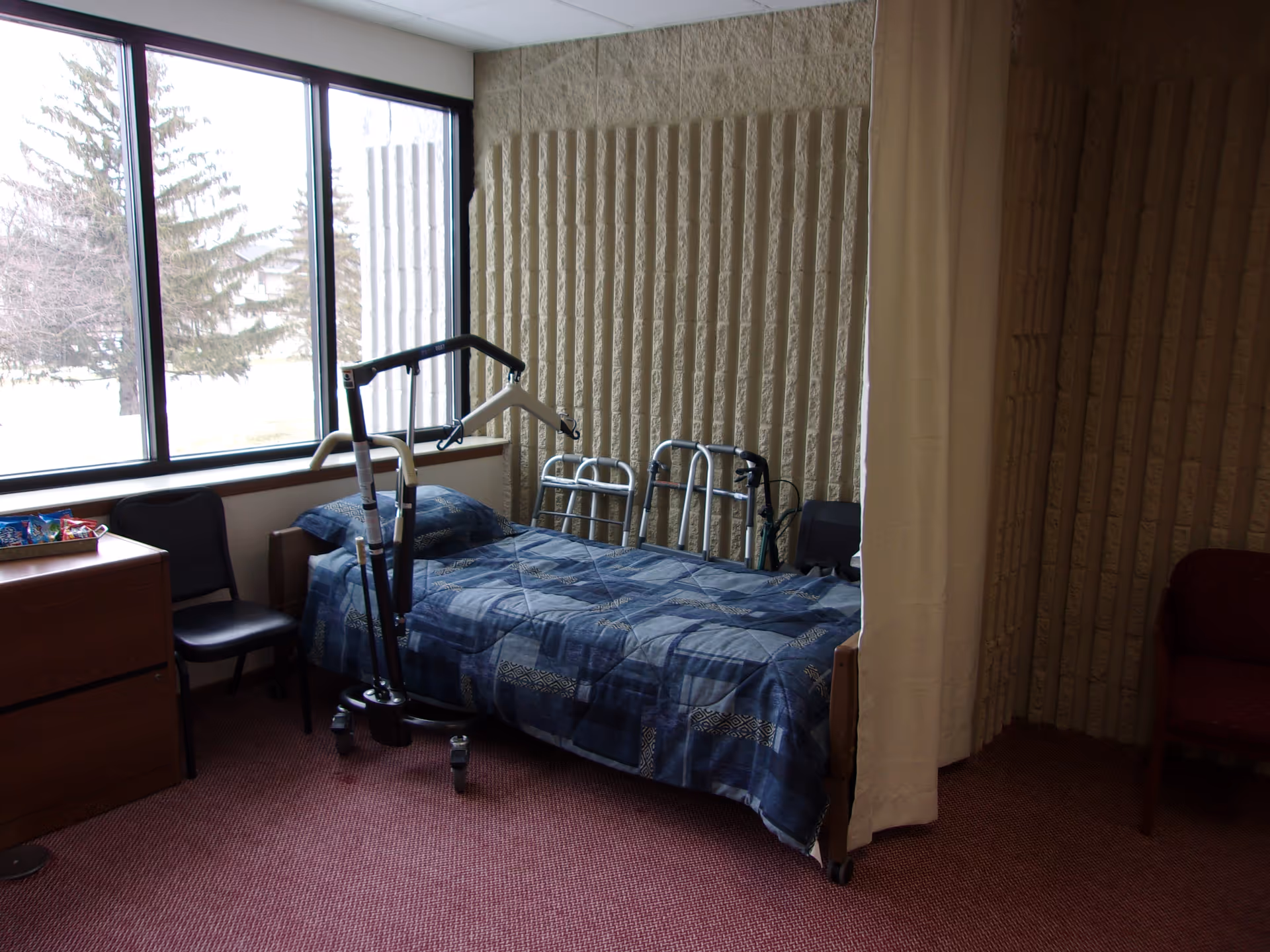 A senior living bedroom with a blue quilted bed, mobility aid, dresser, and large windows.