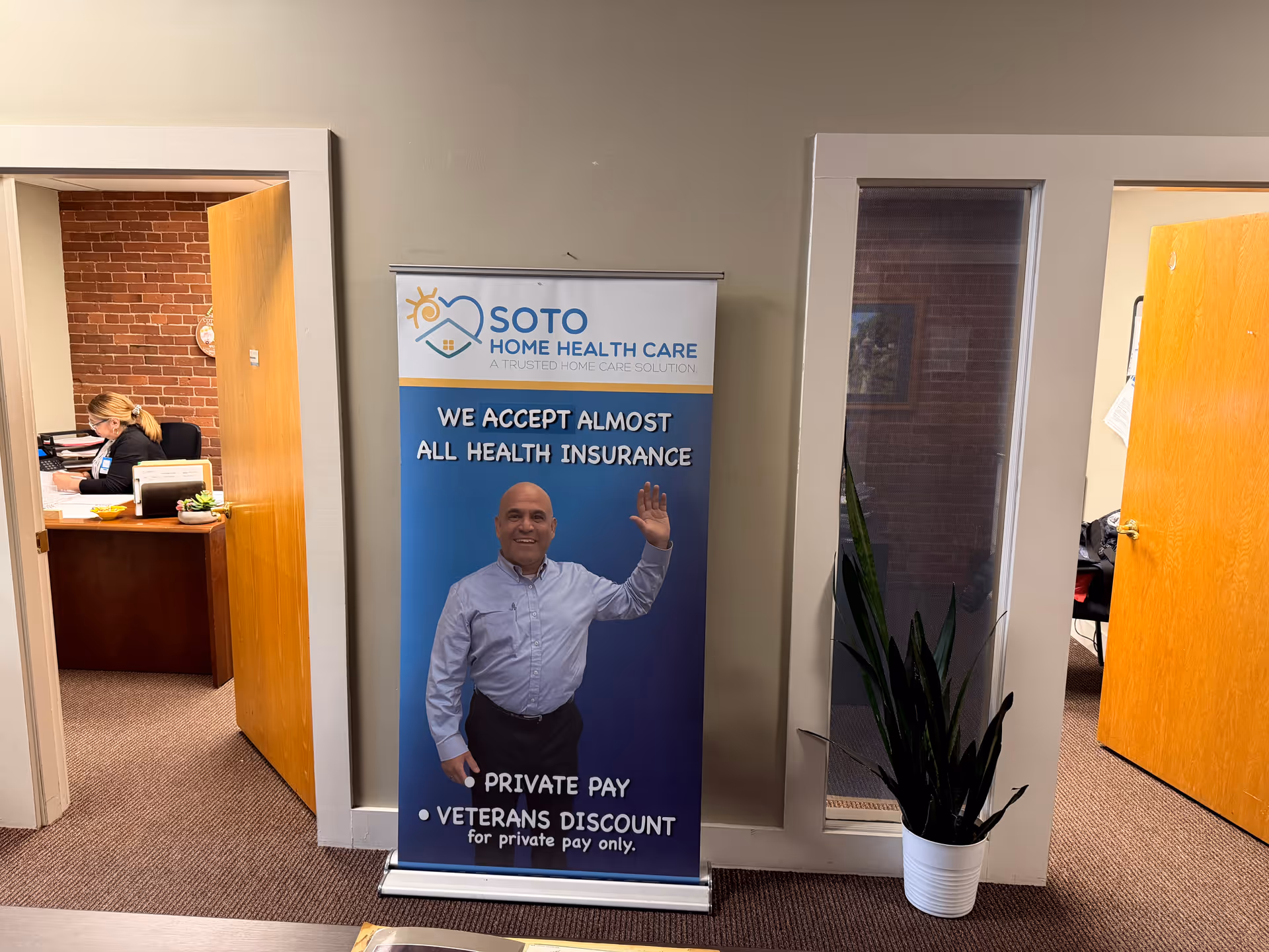 Office hallway with a Soto Home Health Care banner, a desk worker, and a potted plant.