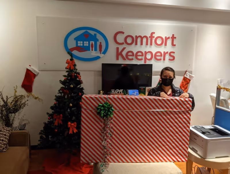 Reception area with staff behind a striped red-and-white desk, Christmas tree, and Comfort Keepers sign.