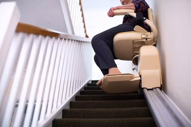 Senior individual seated on a beige stairlift chair along an indoor hallway staircase.