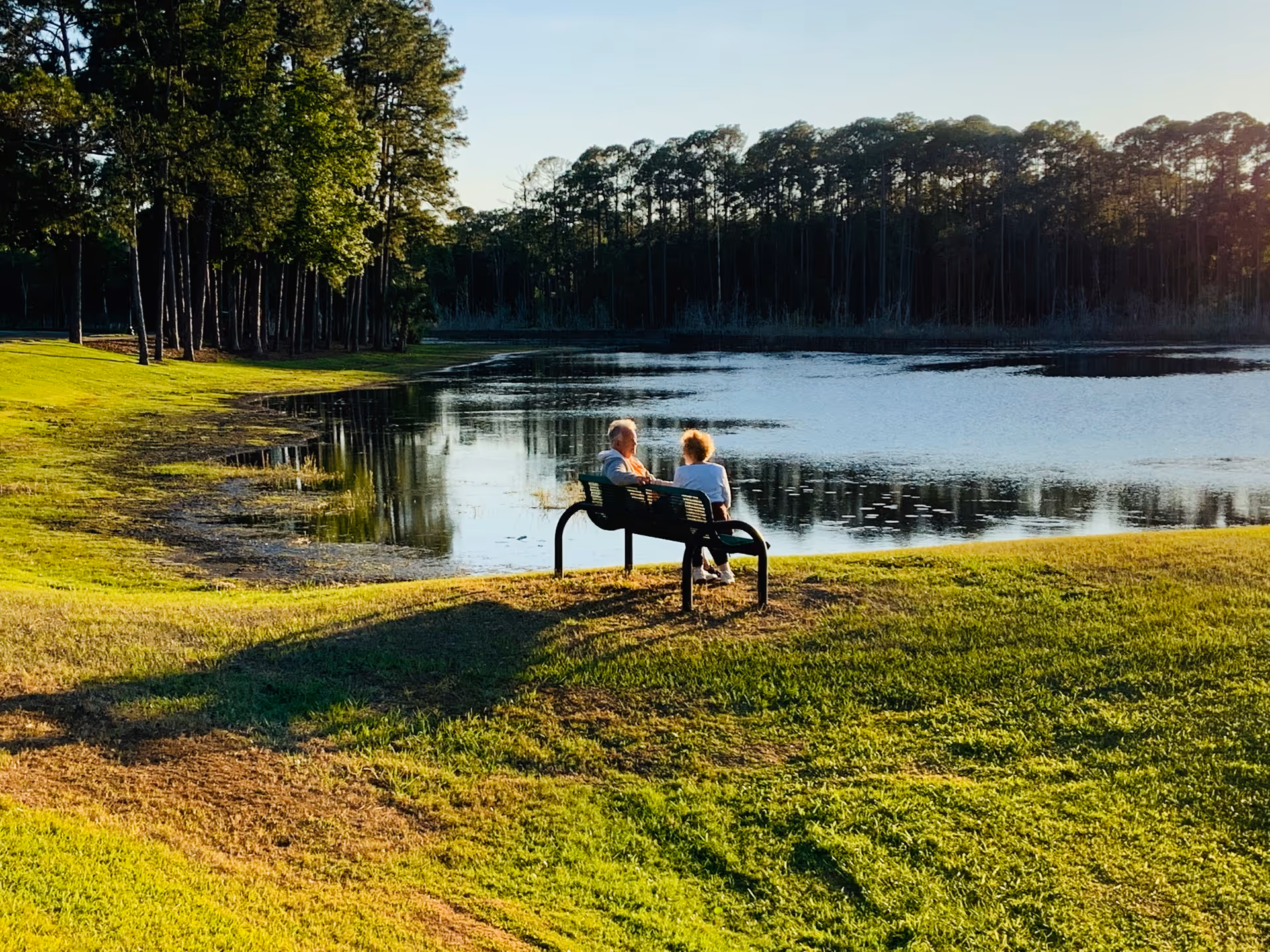 Two elderly people sit on a bench by a lake in a park.