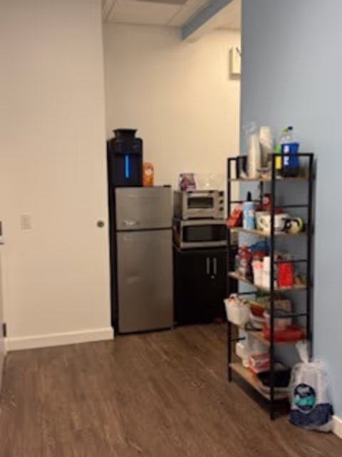 Small kitchenette with refrigerator, microwave, and open shelving stocked with dishes and supplies.