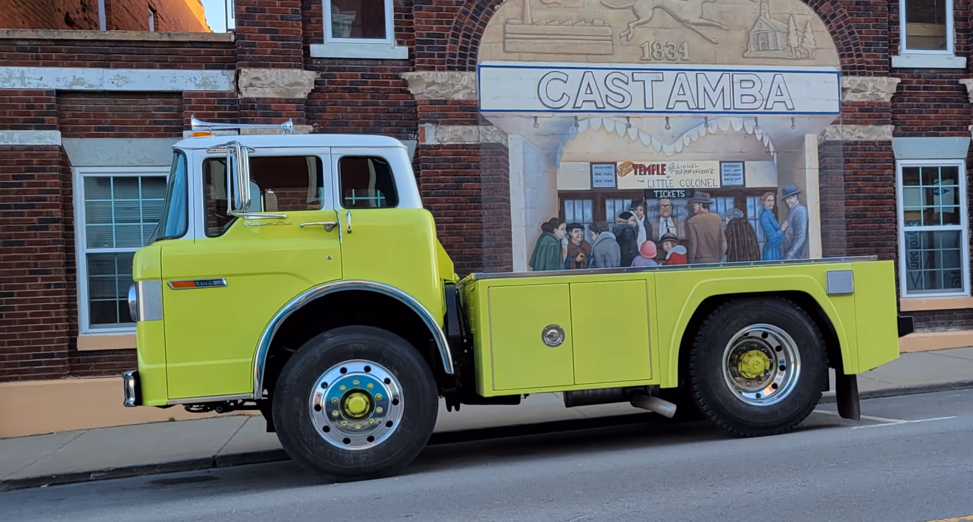 Bright lime-yellow vintage tow truck parked on street in front of a brick building with mural.