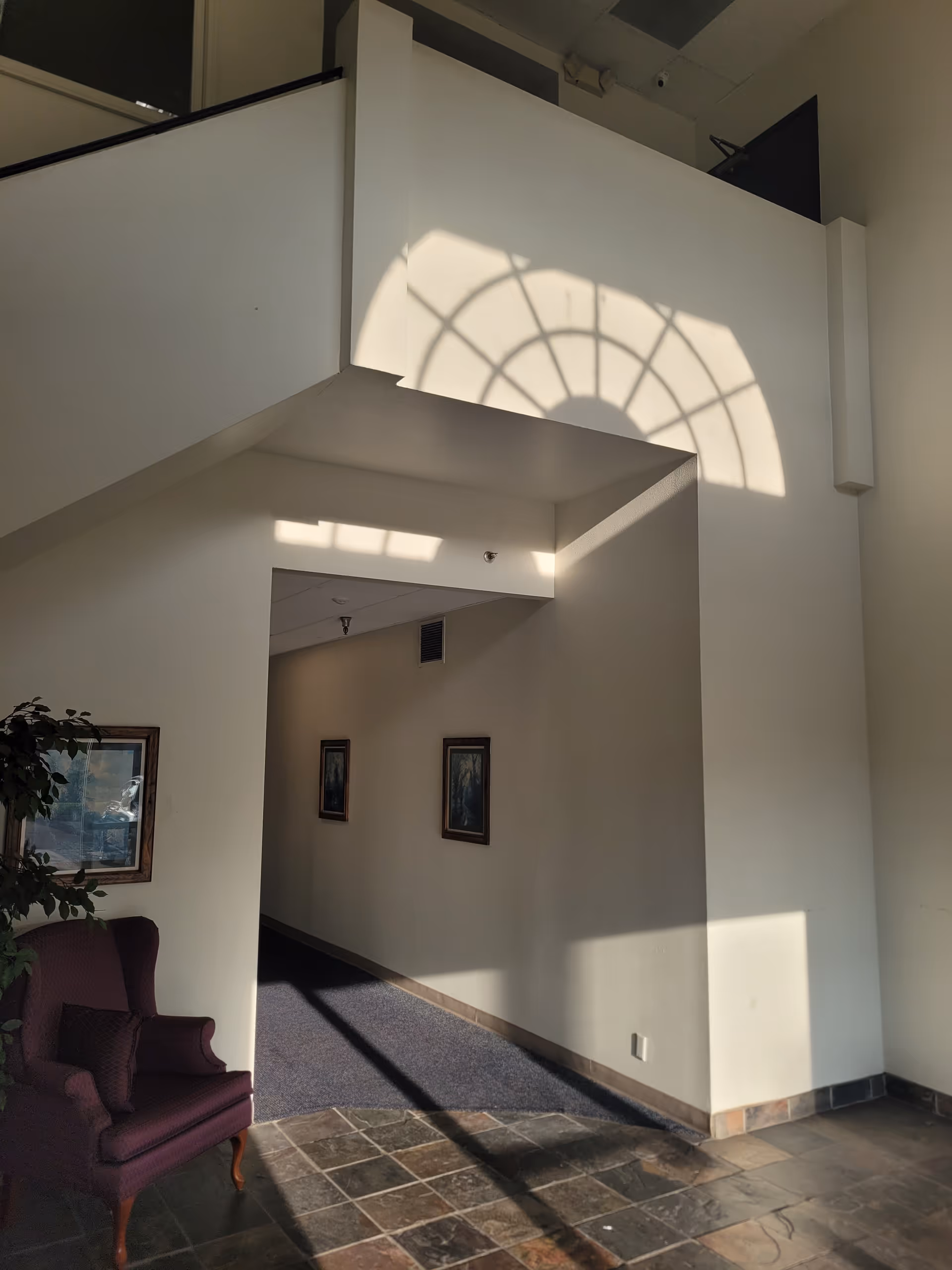 Bright interior hallway with white walls, a purple chair, framed art, and arched shadow.