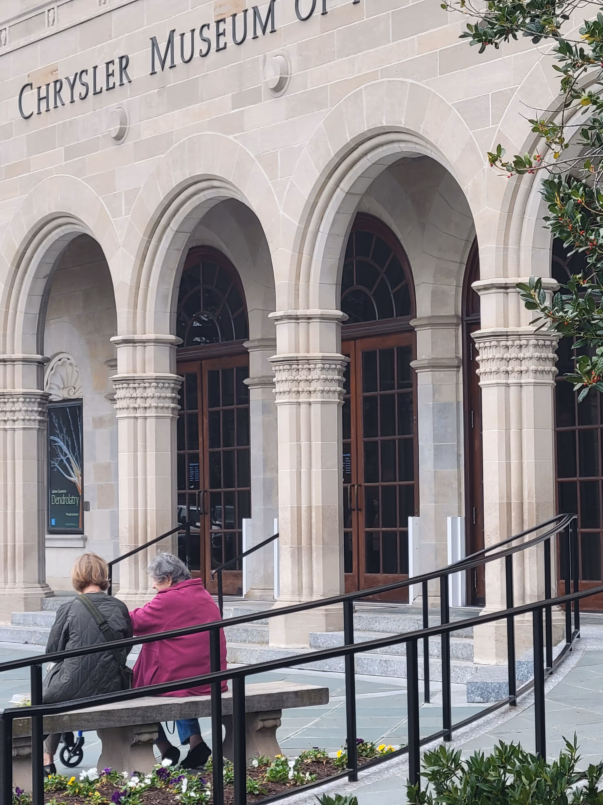 Two elderly people sit on a bench outside the Chrysler Museum of Art entrance.