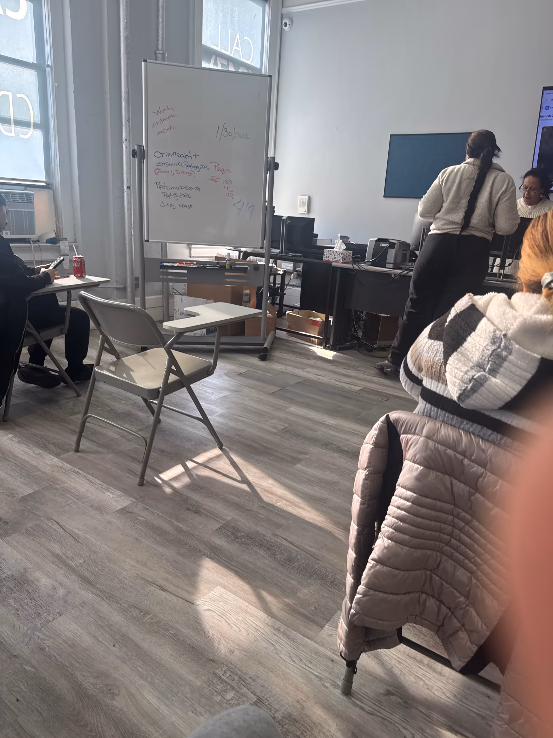 Interior classroom-style room with a whiteboard, desks, chairs, and several people interacting.