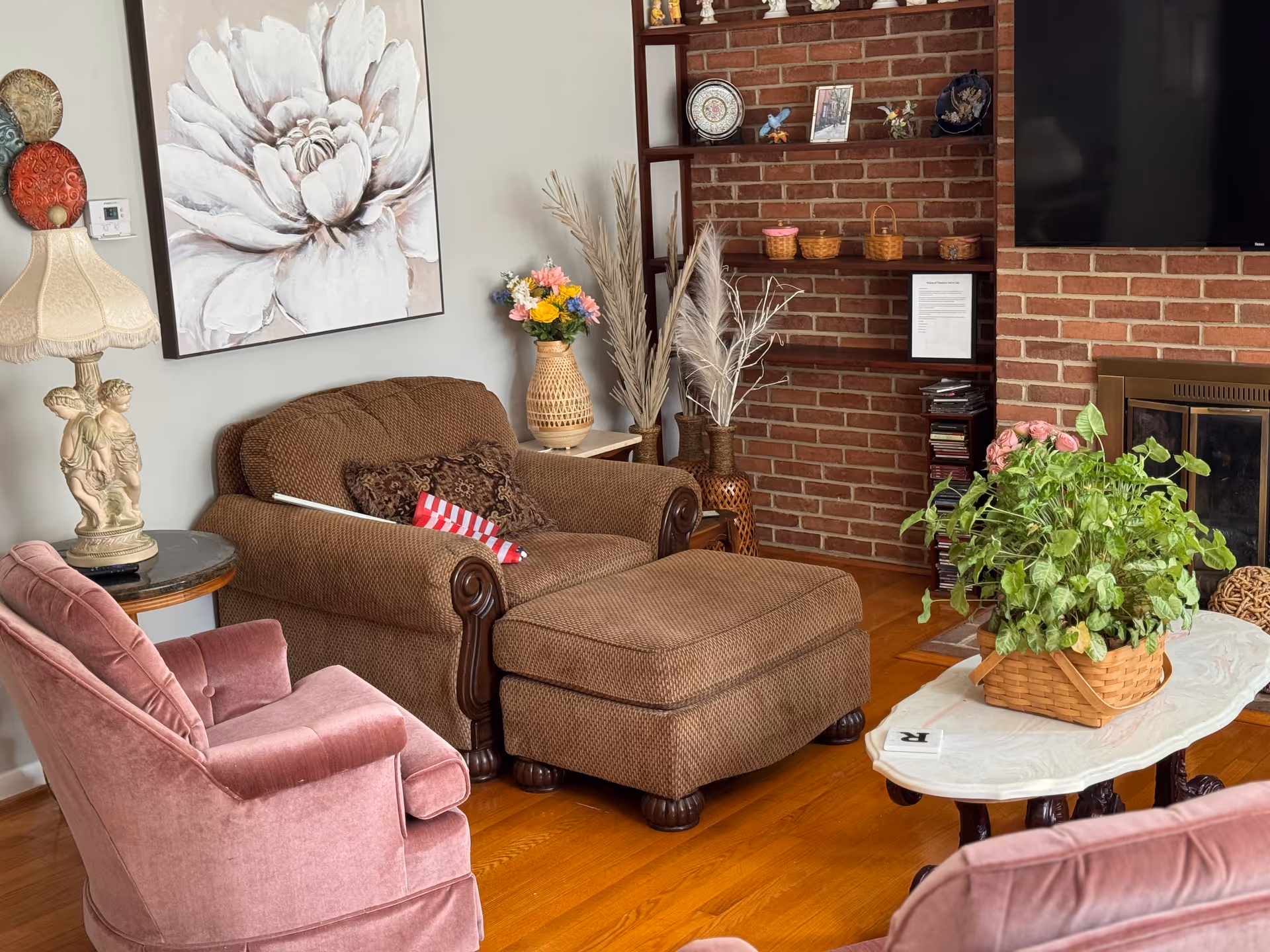 Cozy living room featuring pink chairs, brown sofa, brick fireplace, shelves, and plants.