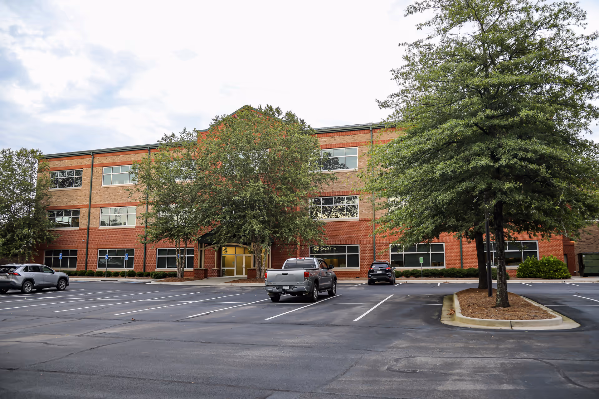 Brick three-story building with a parking lot and trees in front; main entrance visible.