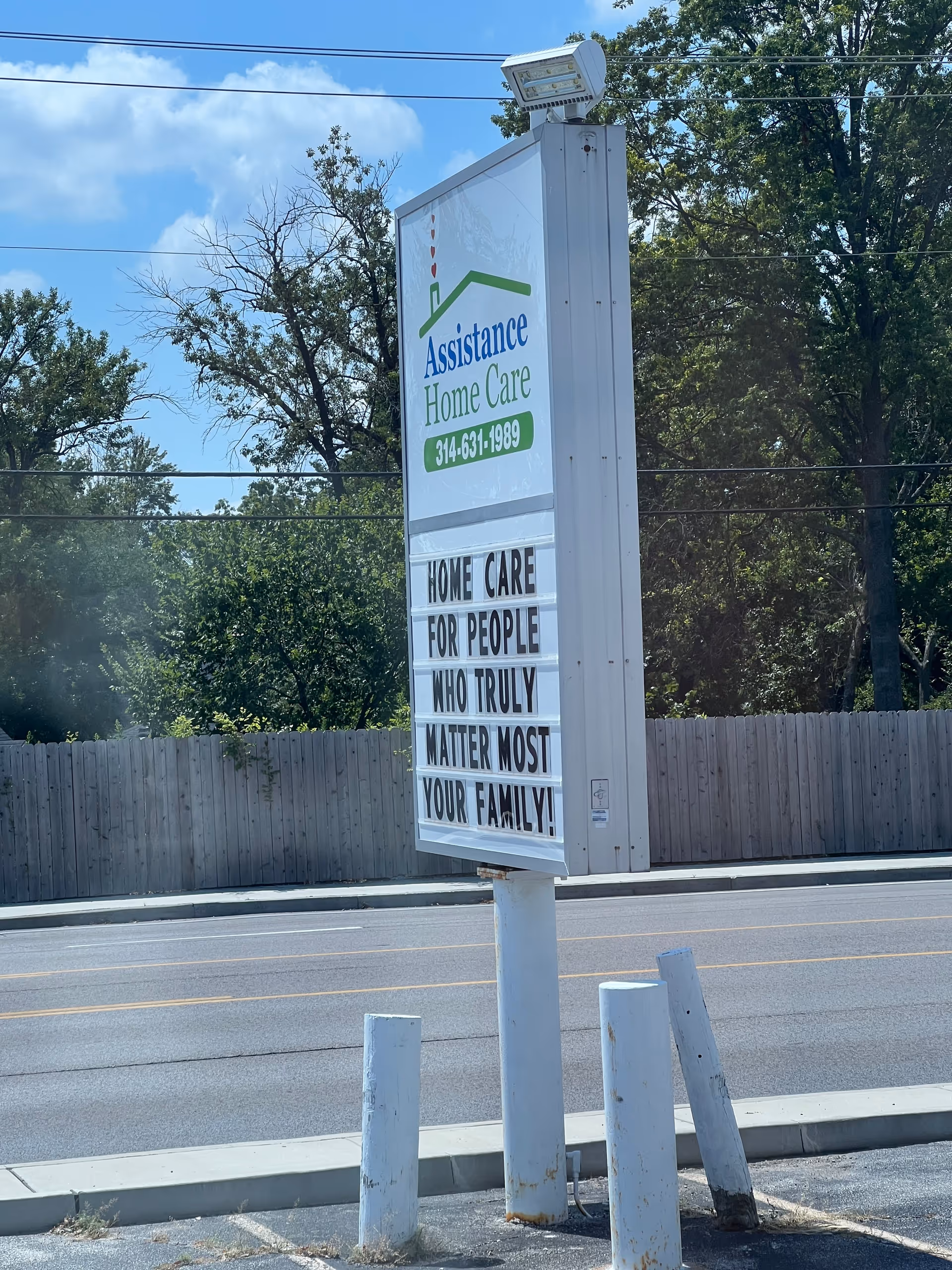 Roadside sign for Assistance Home Care beside a fence, road, and trees.