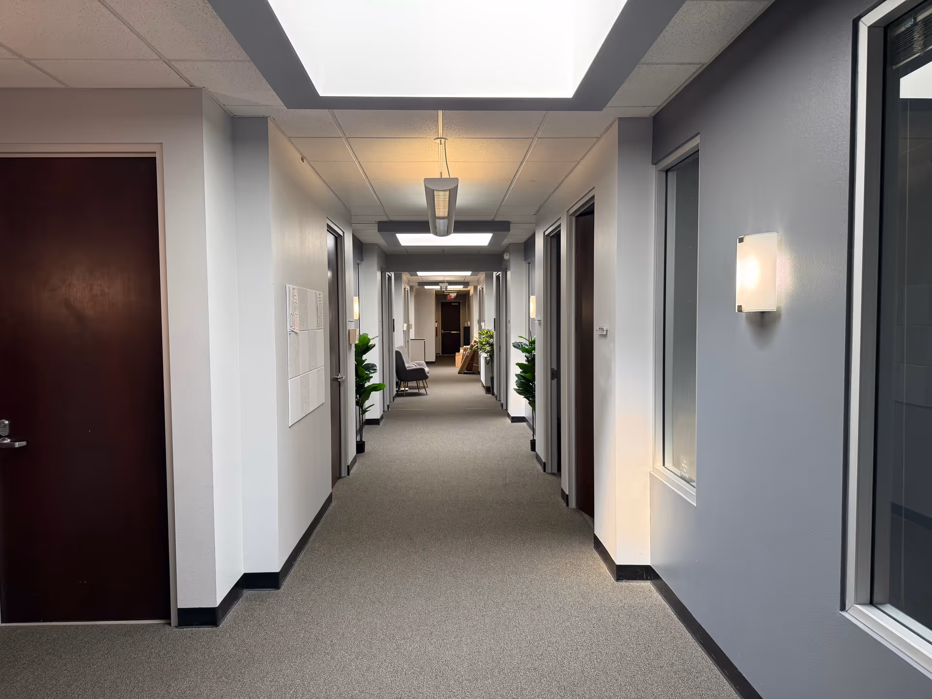 Well-lit interior corridor of a senior living facility with doors, lights, and potted plants.