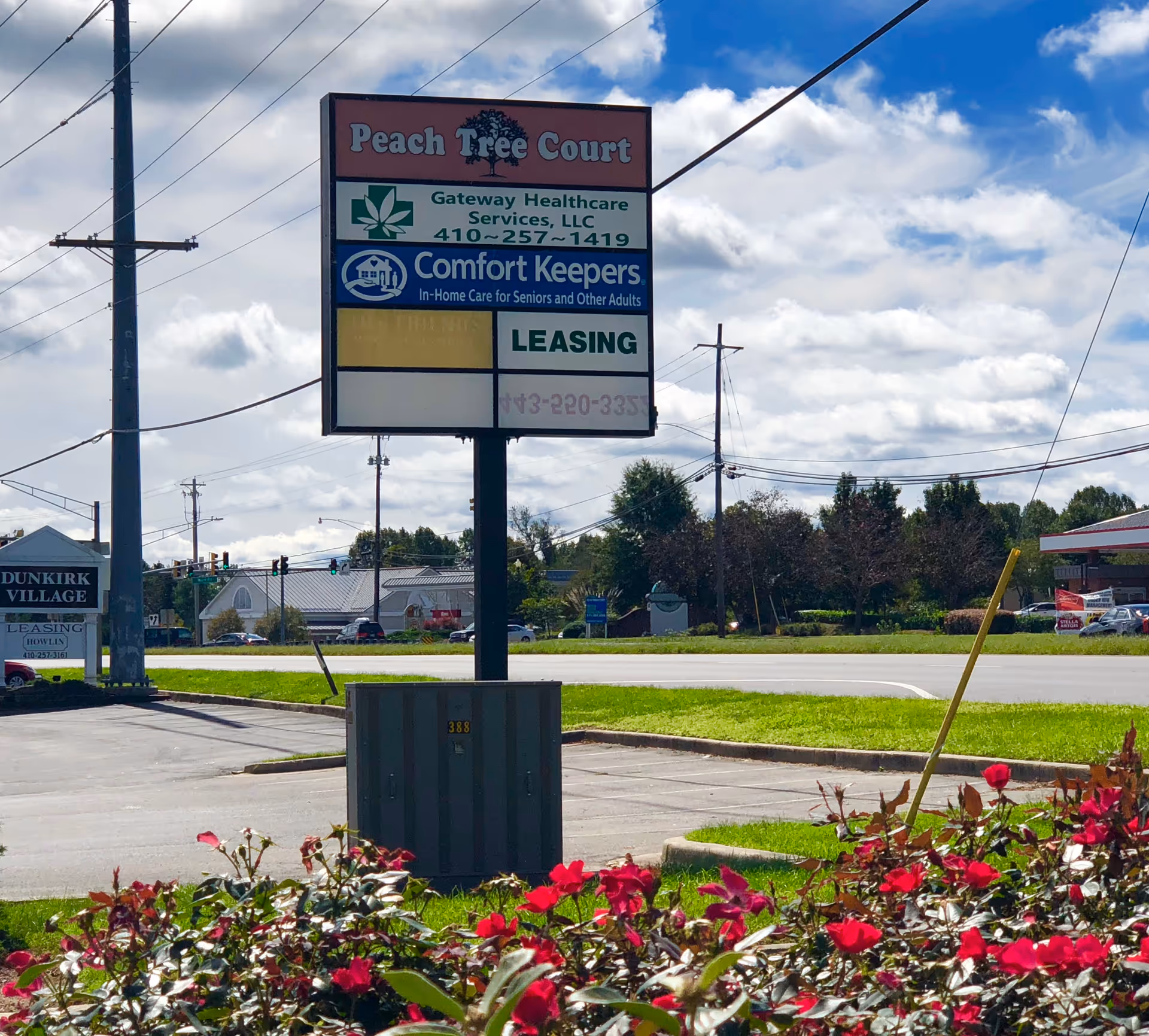 Tall multi-sign board advertising Peach Tree Court and Comfort Keepers, with a parking lot and flowers.