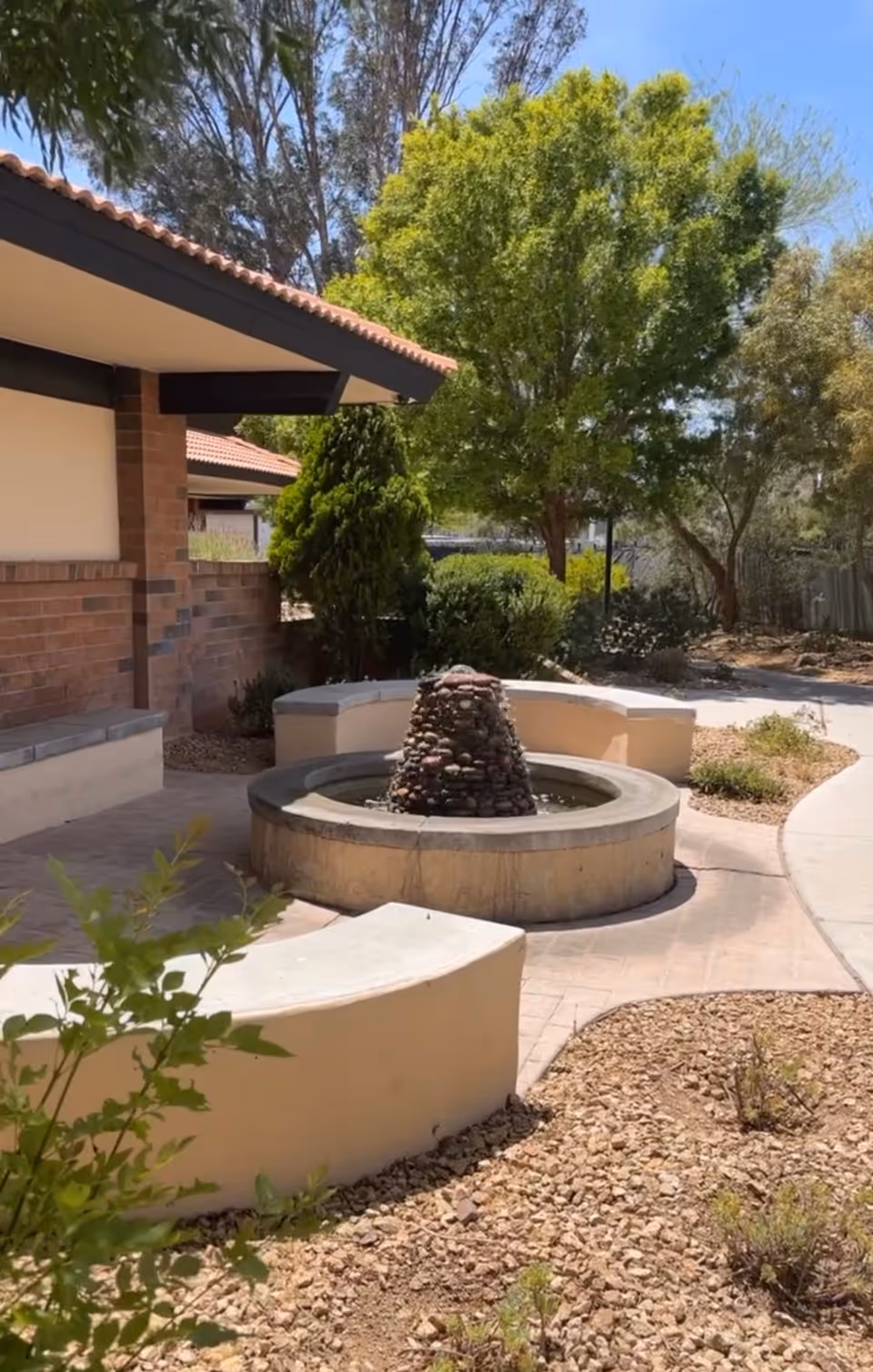 Outdoor courtyard with a circular stone fountain and curved seating beside a brick building.