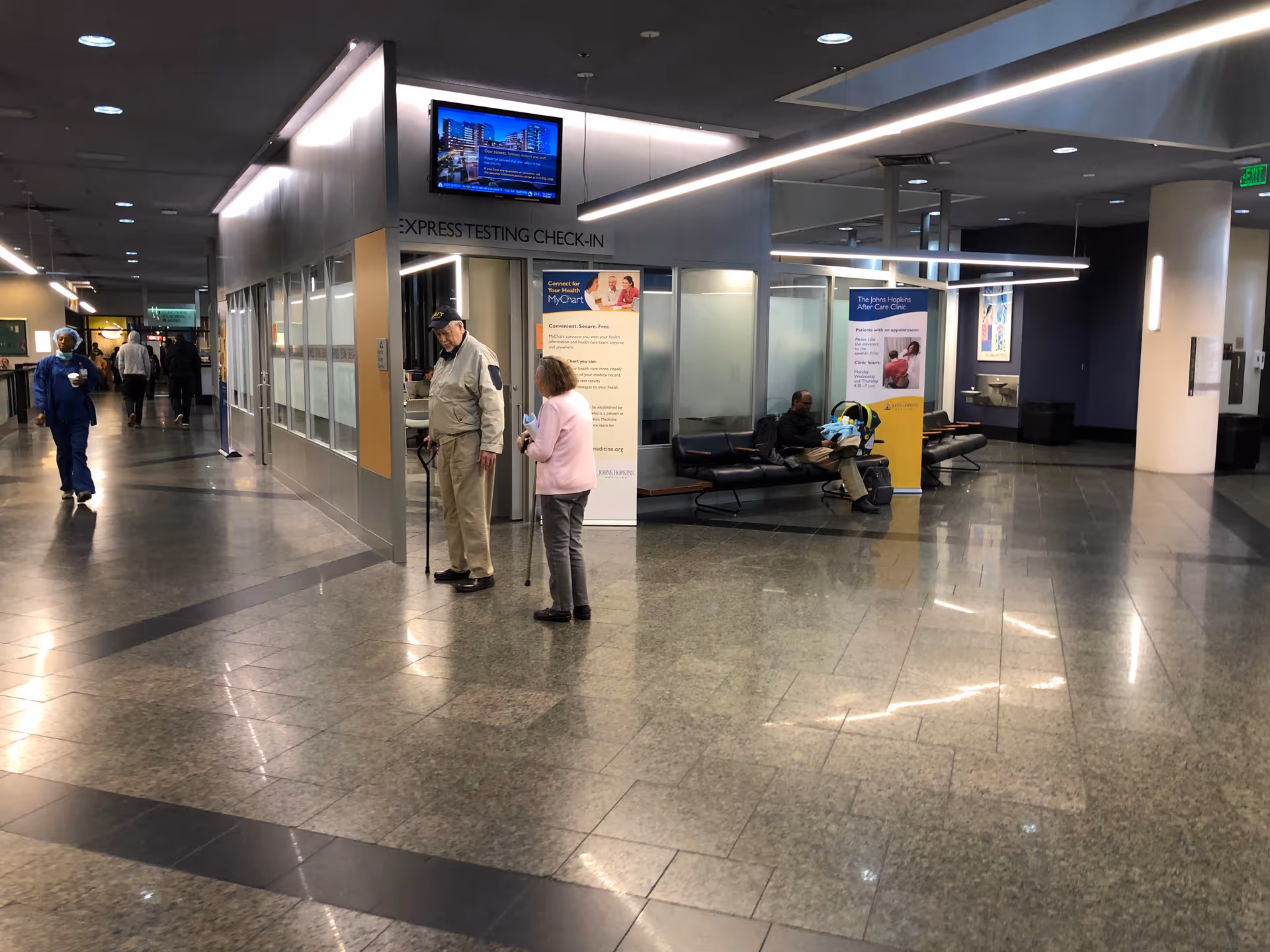 Hospital corridor with check-in area, benches, and informational posters in a medical center.