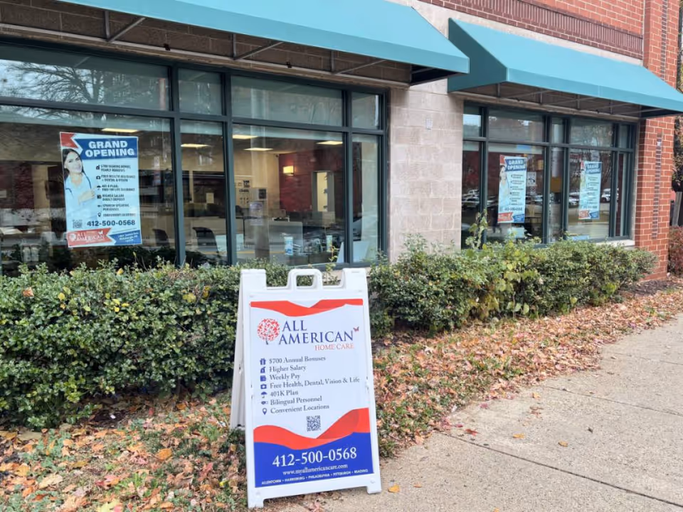 Exterior view of All American Home Care storefront with posters and a sidewalk sign.