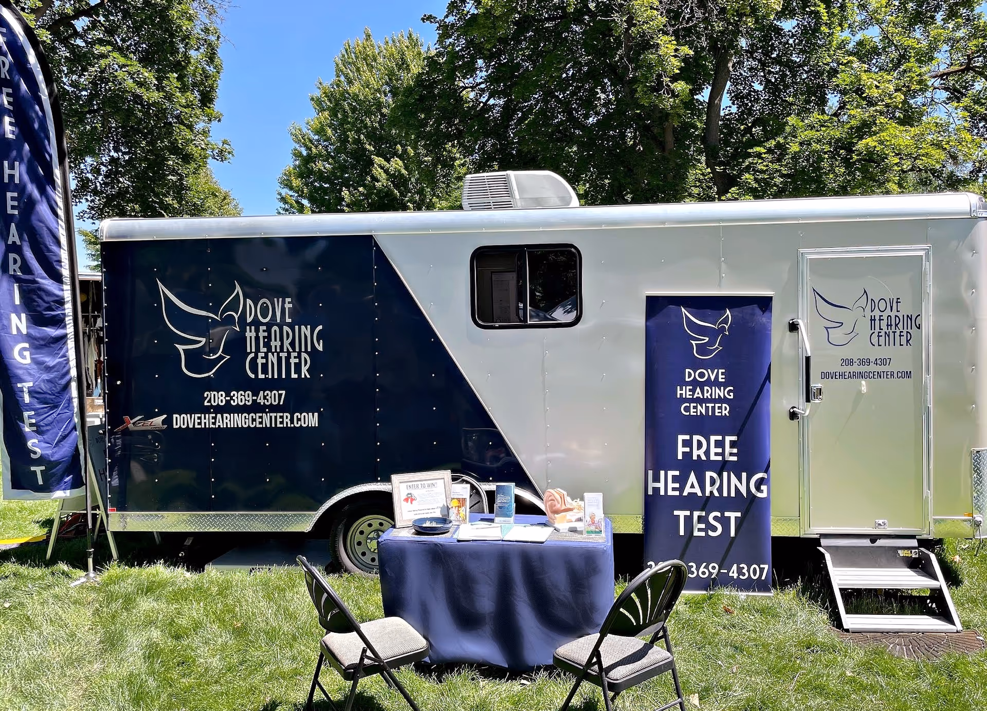 Outdoor mobile hearing center trailer with banners, table, and chairs on grassy area.
