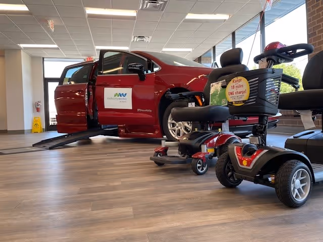 Red car on a lift inside a showroom, with mobility scooter in the foreground.