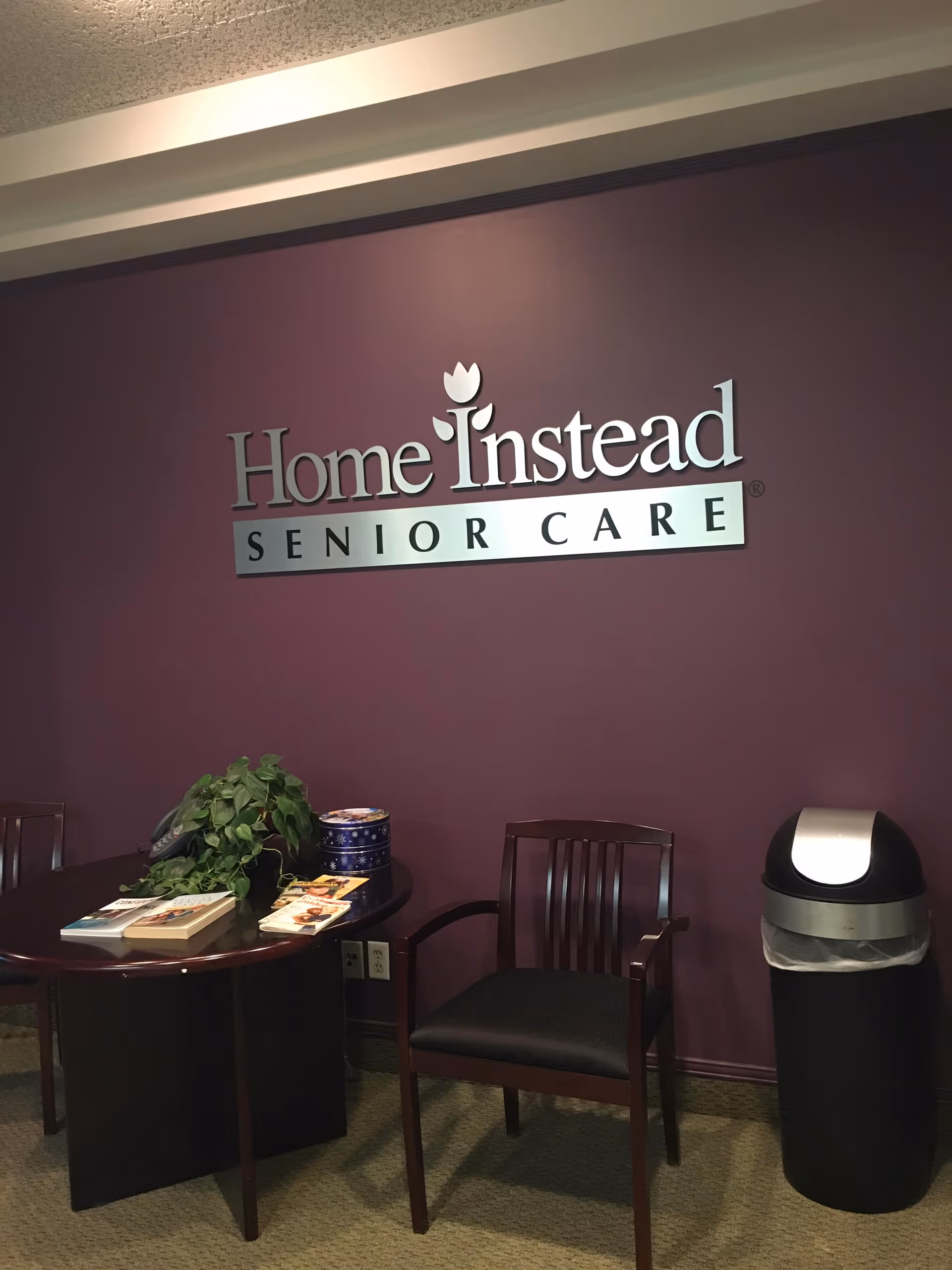 Interior reception area with purple wall, Home Instead Senior Care logo, chair, table, and trash can.