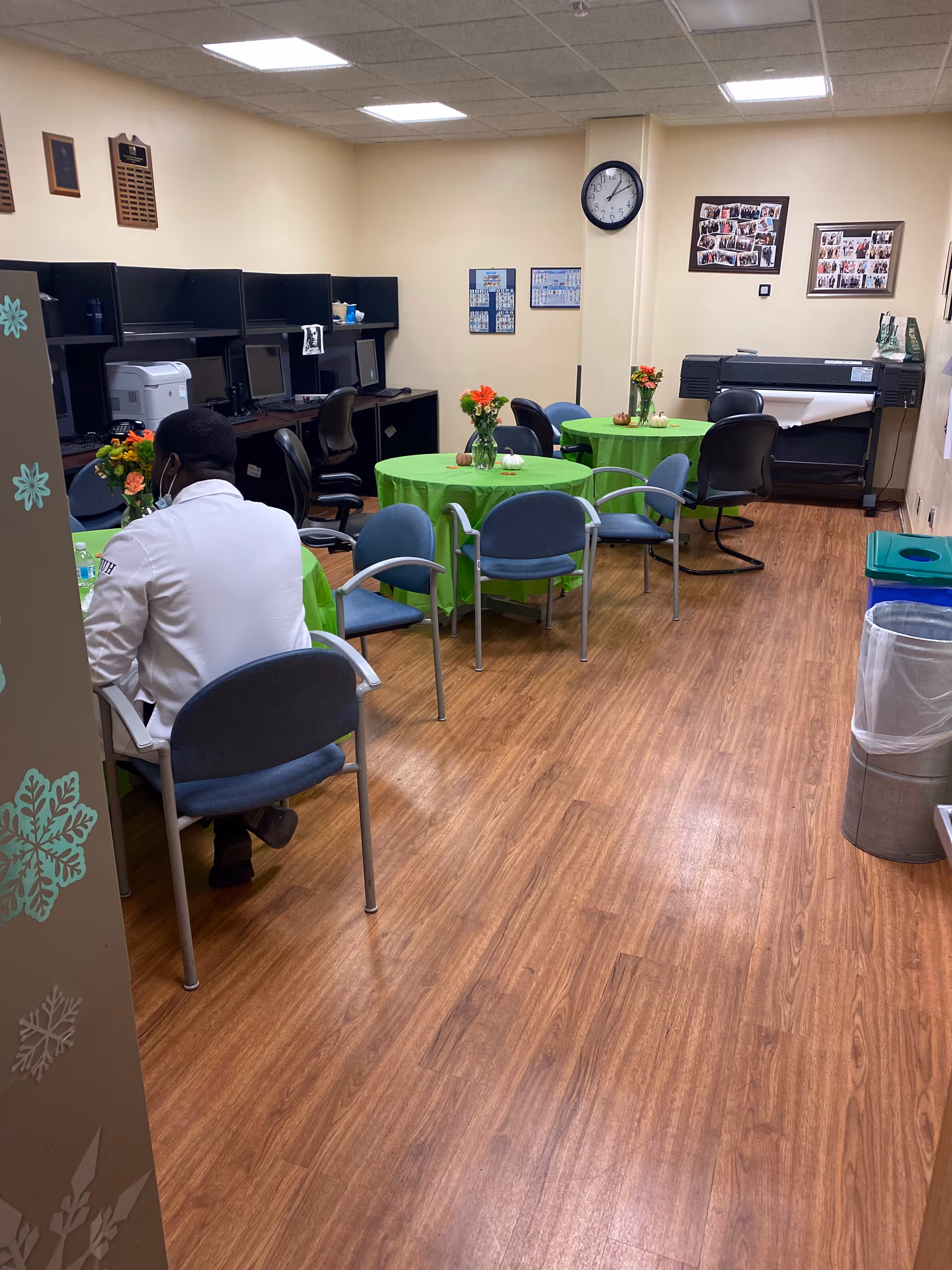Indoor facility lounge with green-tablecloth tables, chairs, a clock, and a staff member