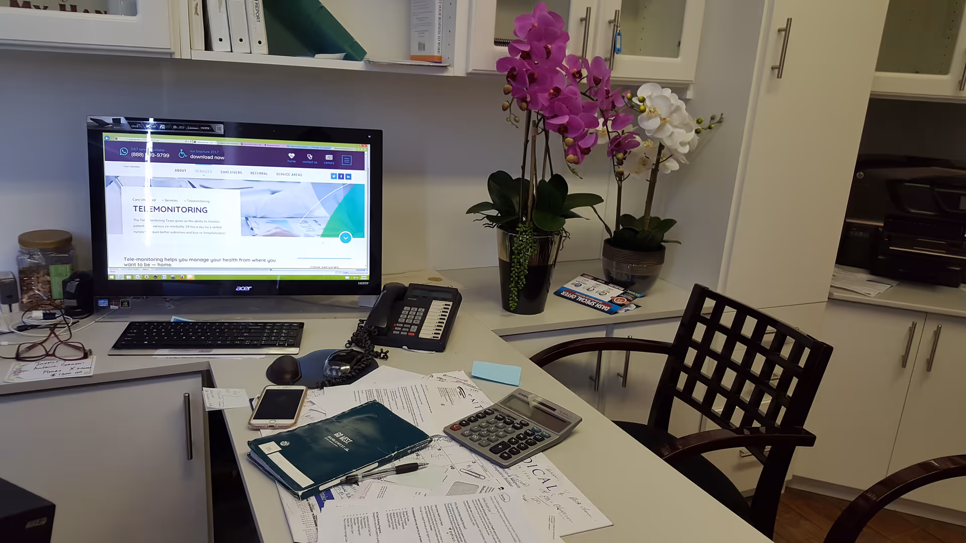 Office desk at Care Unlimited Health Services Inc with computer, phone, papers, and orchids.