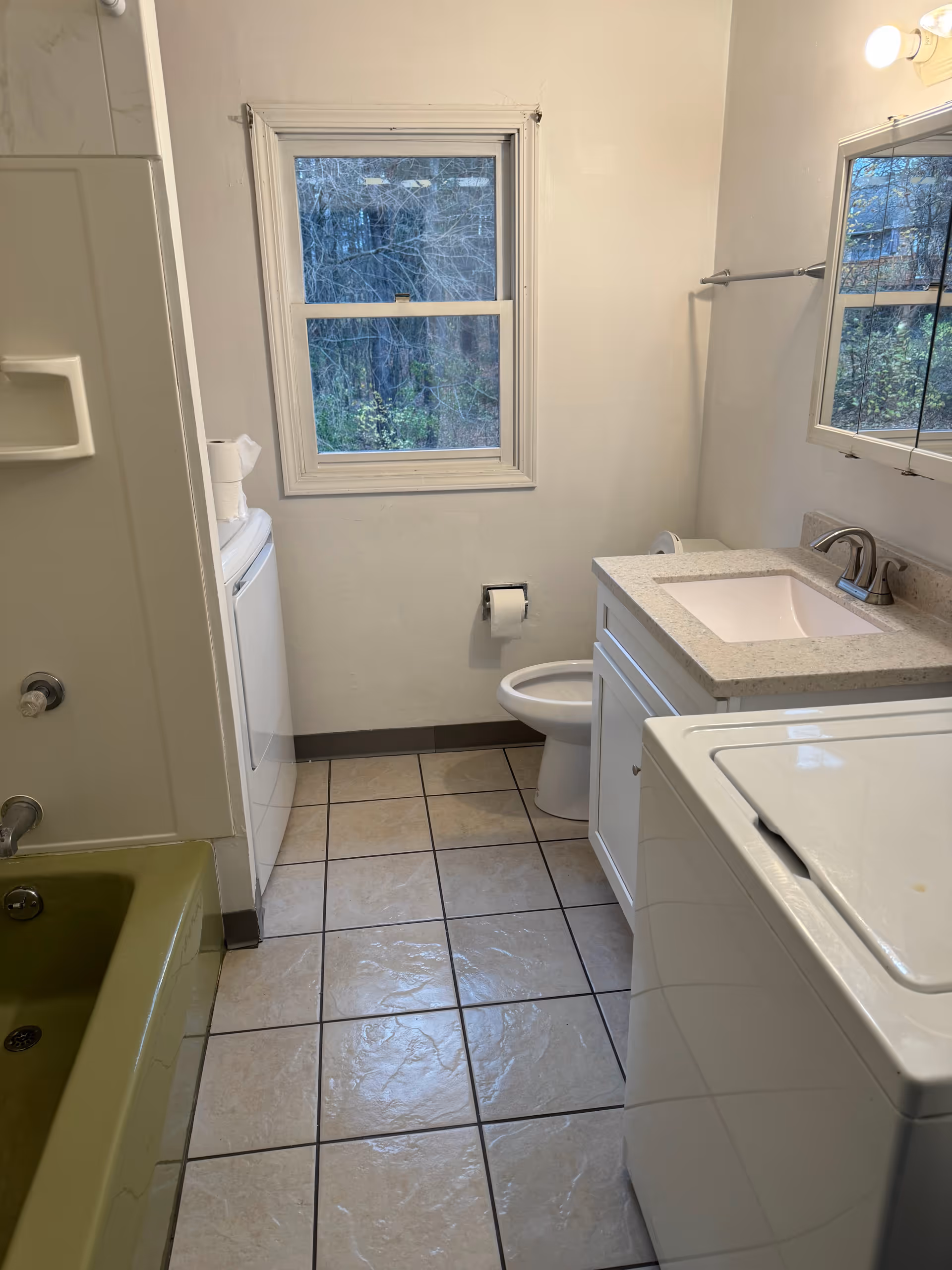 Small shared bathroom with sink, toilet, washing machine, window, mirror, and green tub.